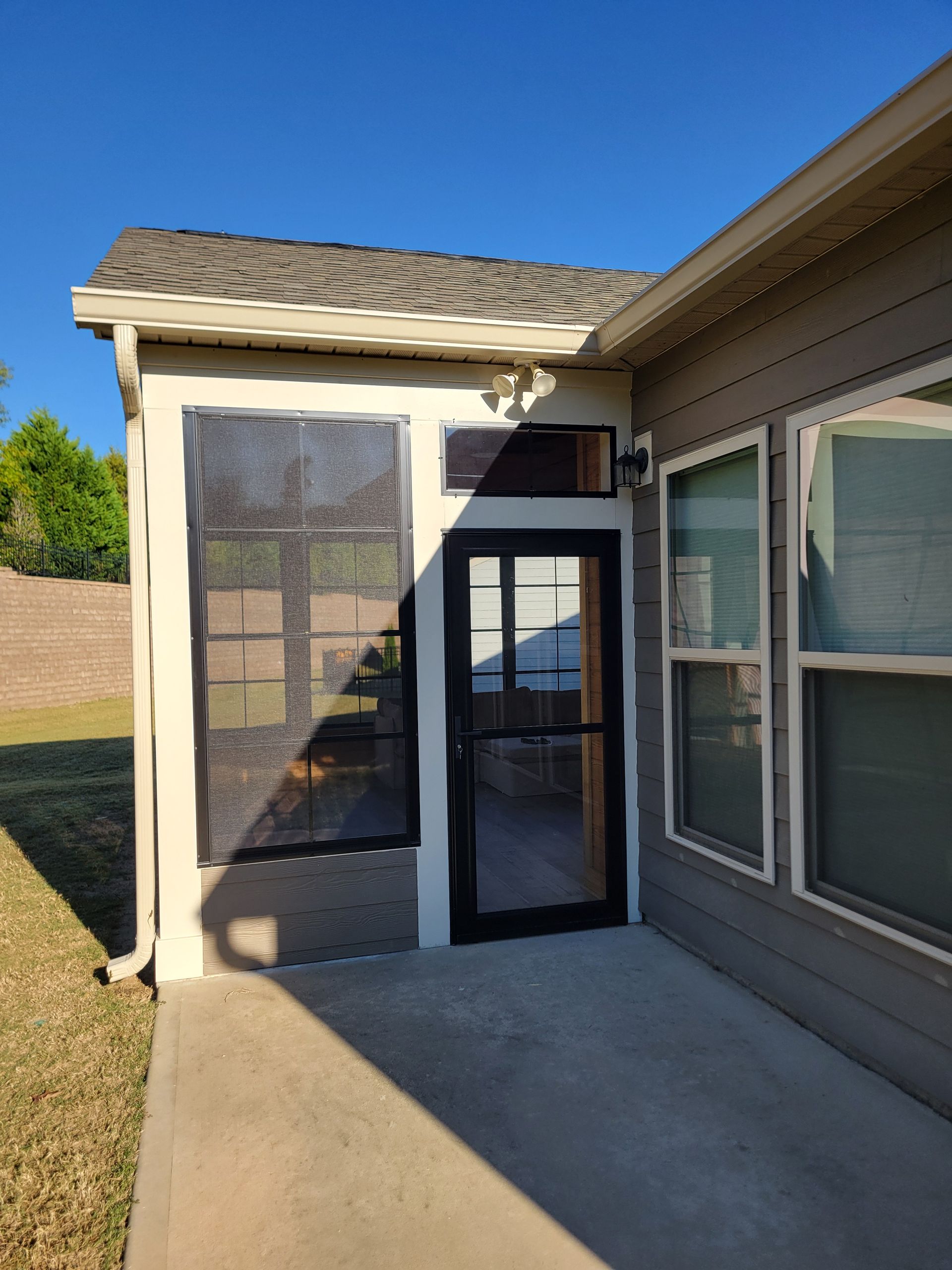 Covered patio with screened door and windows beside a house, lit by bright sunlight.