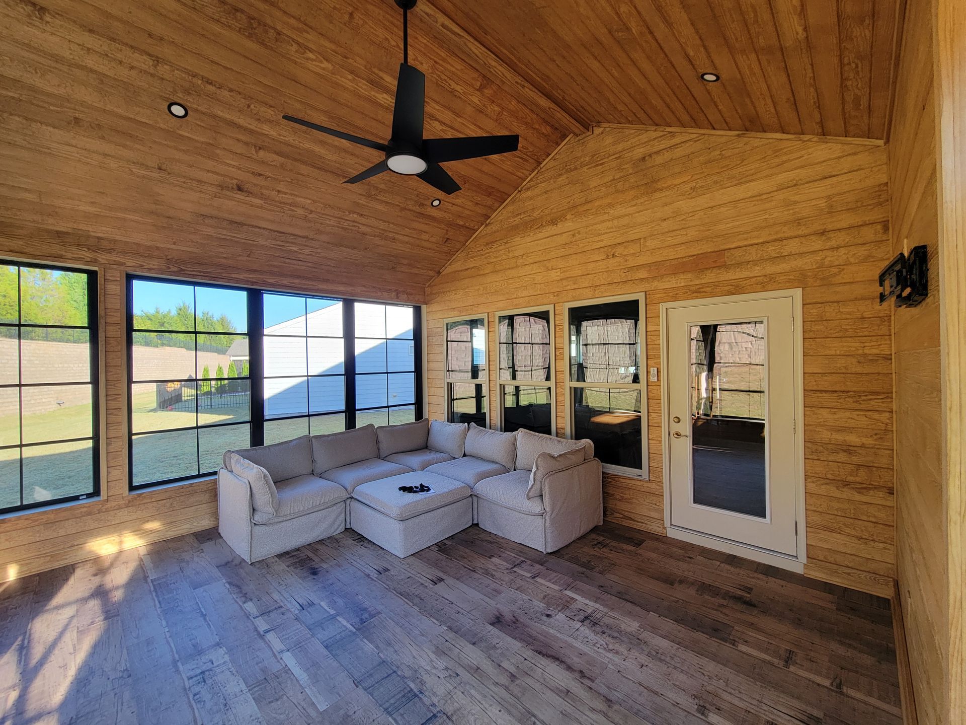 Covered porch with wood-paneled walls, large windows, a ceiling fan, and a light gray sectional sofa.