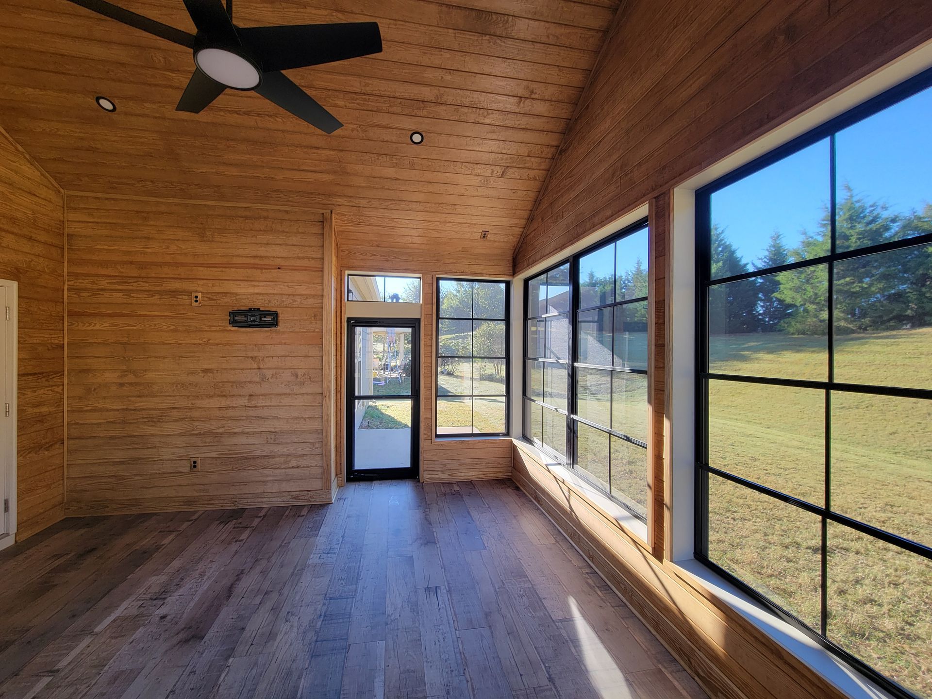 Empty sunroom with wood-paneled walls, vaulted ceiling, ceiling fan, and large windows overlooking a grassy yard