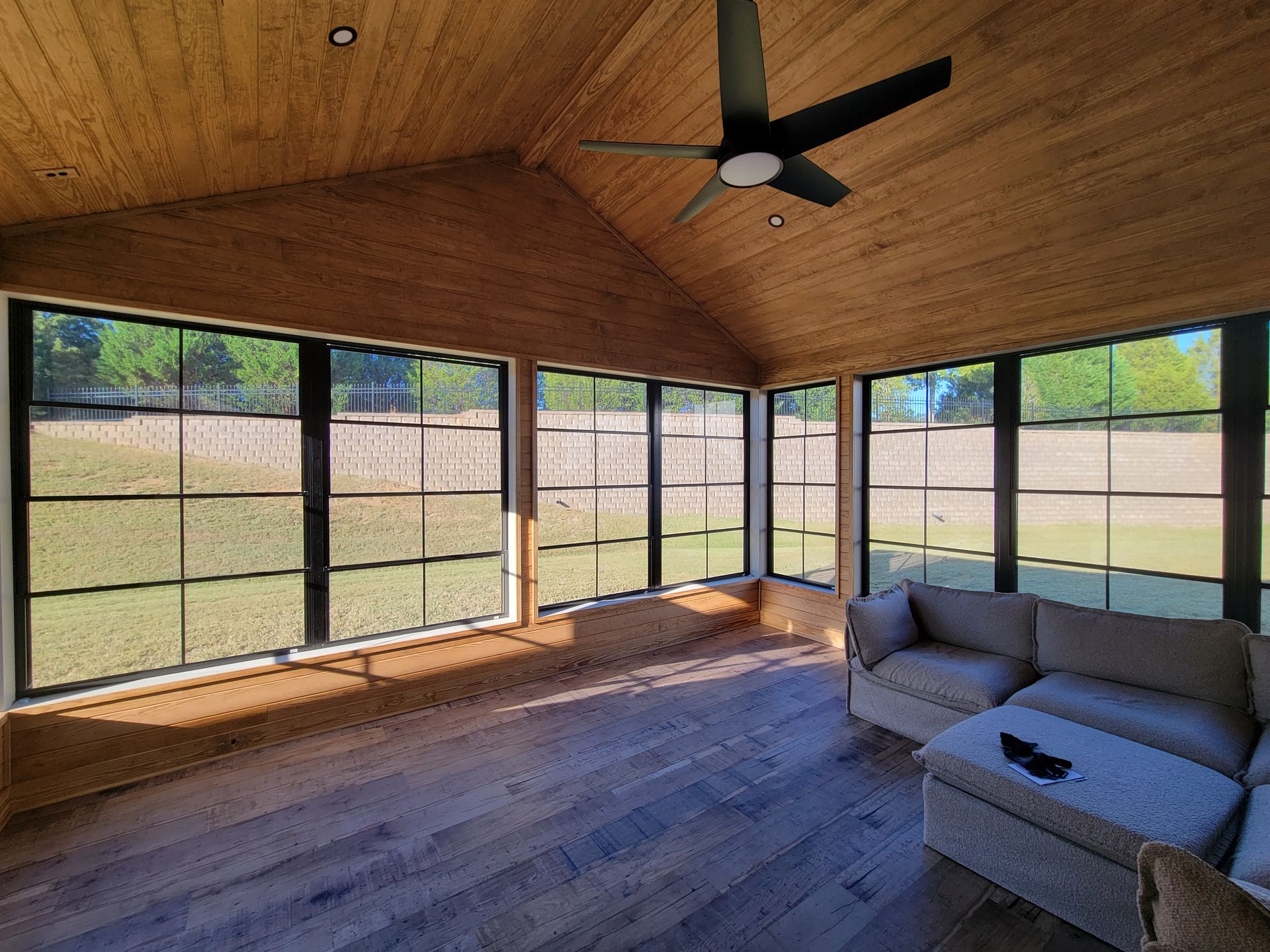 Screened porch with wood ceiling, ceiling fan, gray sectional sofa, and open view of a grassy yard.