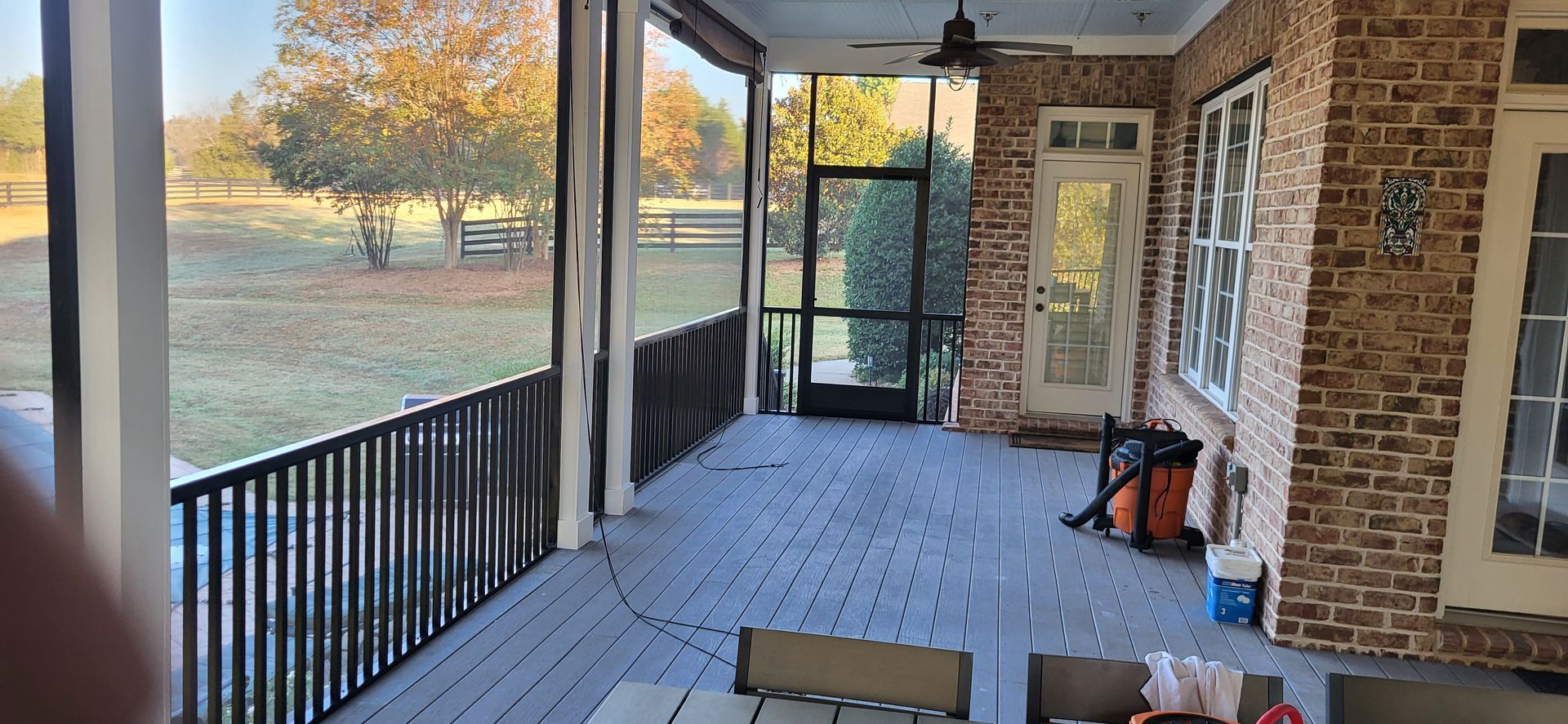 Covered front porch with black railing, brick wall, white columns, and autumn trees beyond the yard.