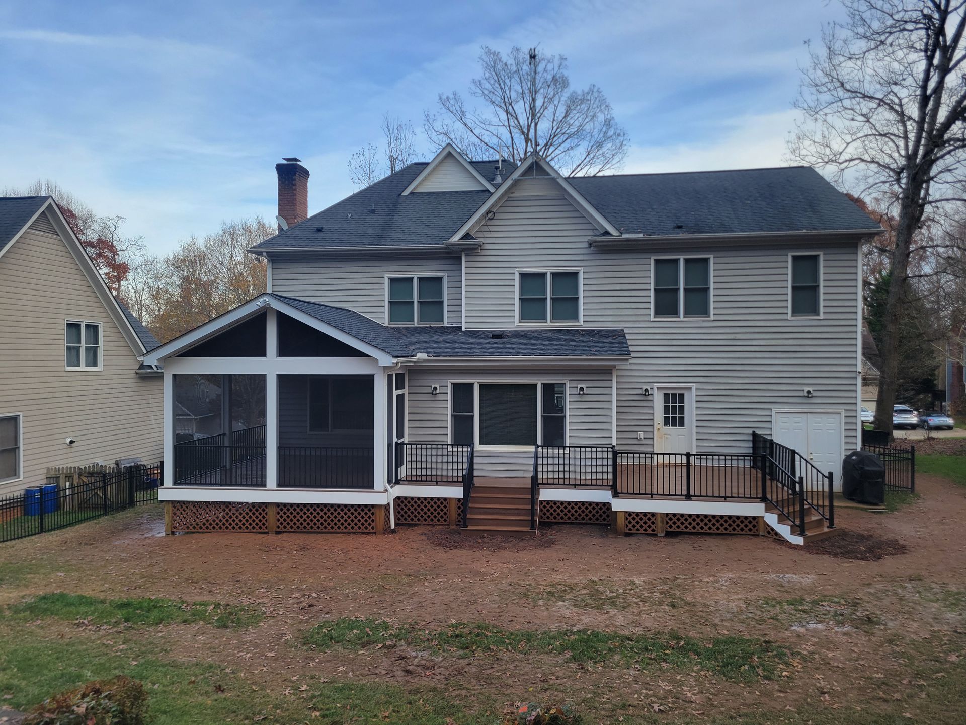Two-story gray house with screened porch, front deck, and leaf-covered yard under a blue sky