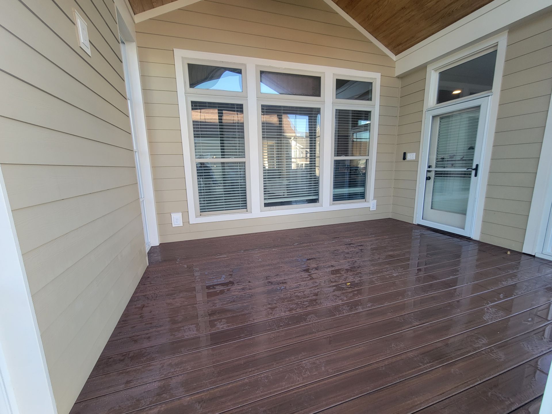 Covered front porch with wood ceiling, brown decking, beige siding, and a glass-paneled door by windows