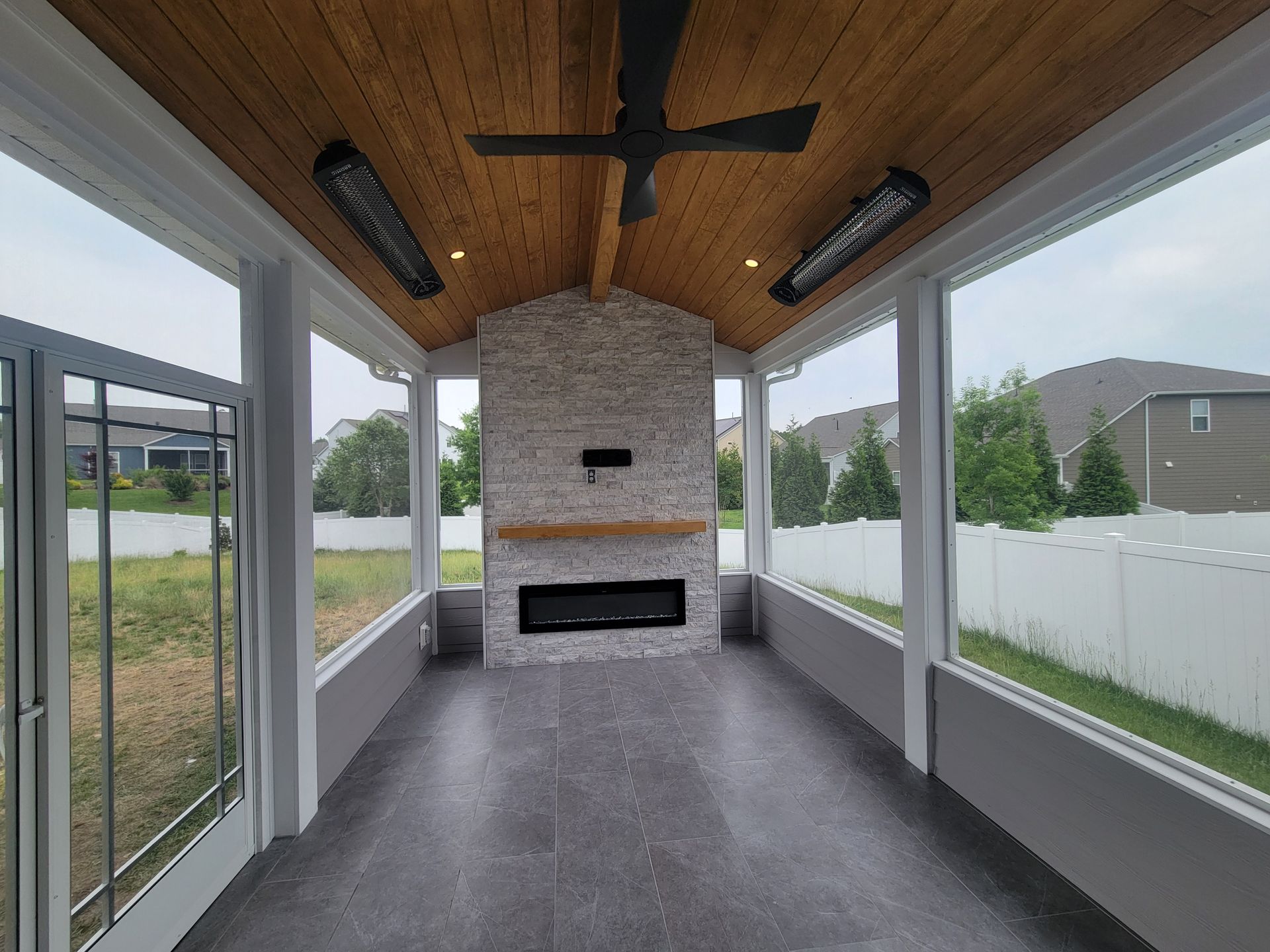 Covered patio with wood ceiling, stone fireplace, and sliding glass doors beside a fenced yard.