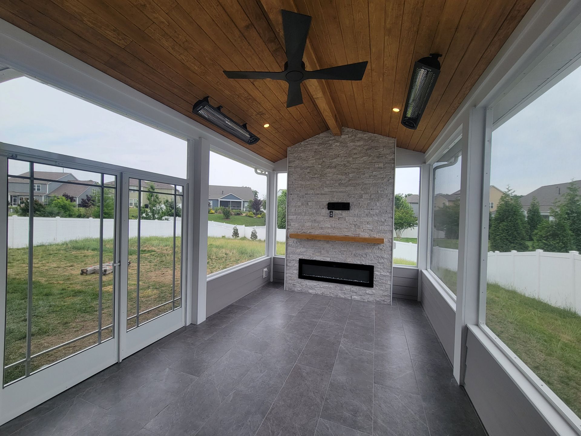 Covered patio with ceiling fan, wood ceiling, stone fireplace, and glass walls overlooking a grassy yard.