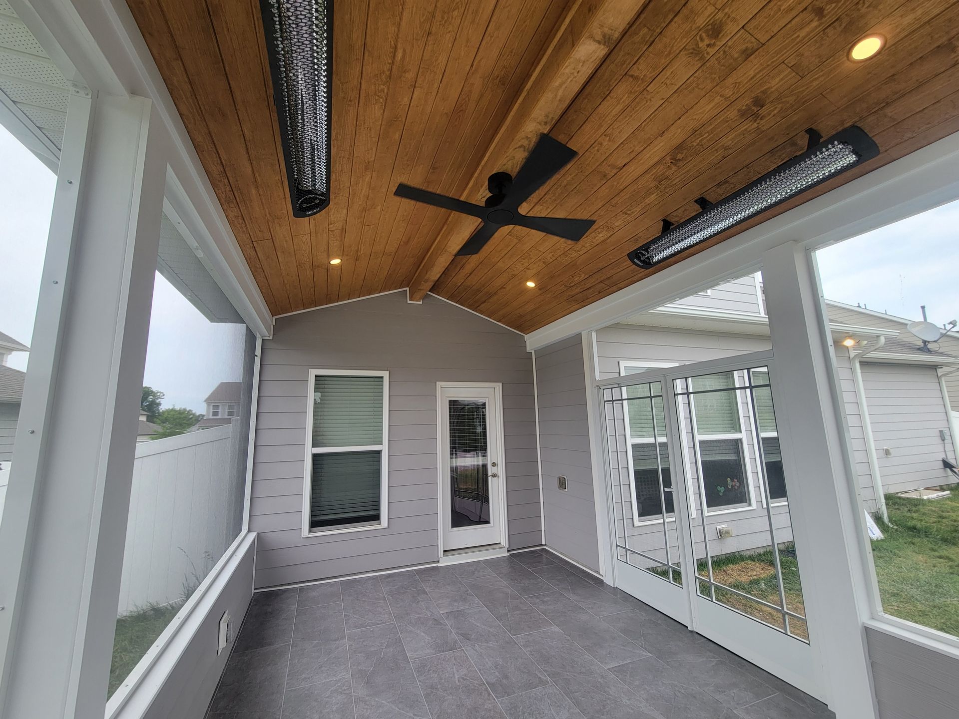 Covered patio with wood ceiling, ceiling fans, and screened windows overlooking a gray backyard.