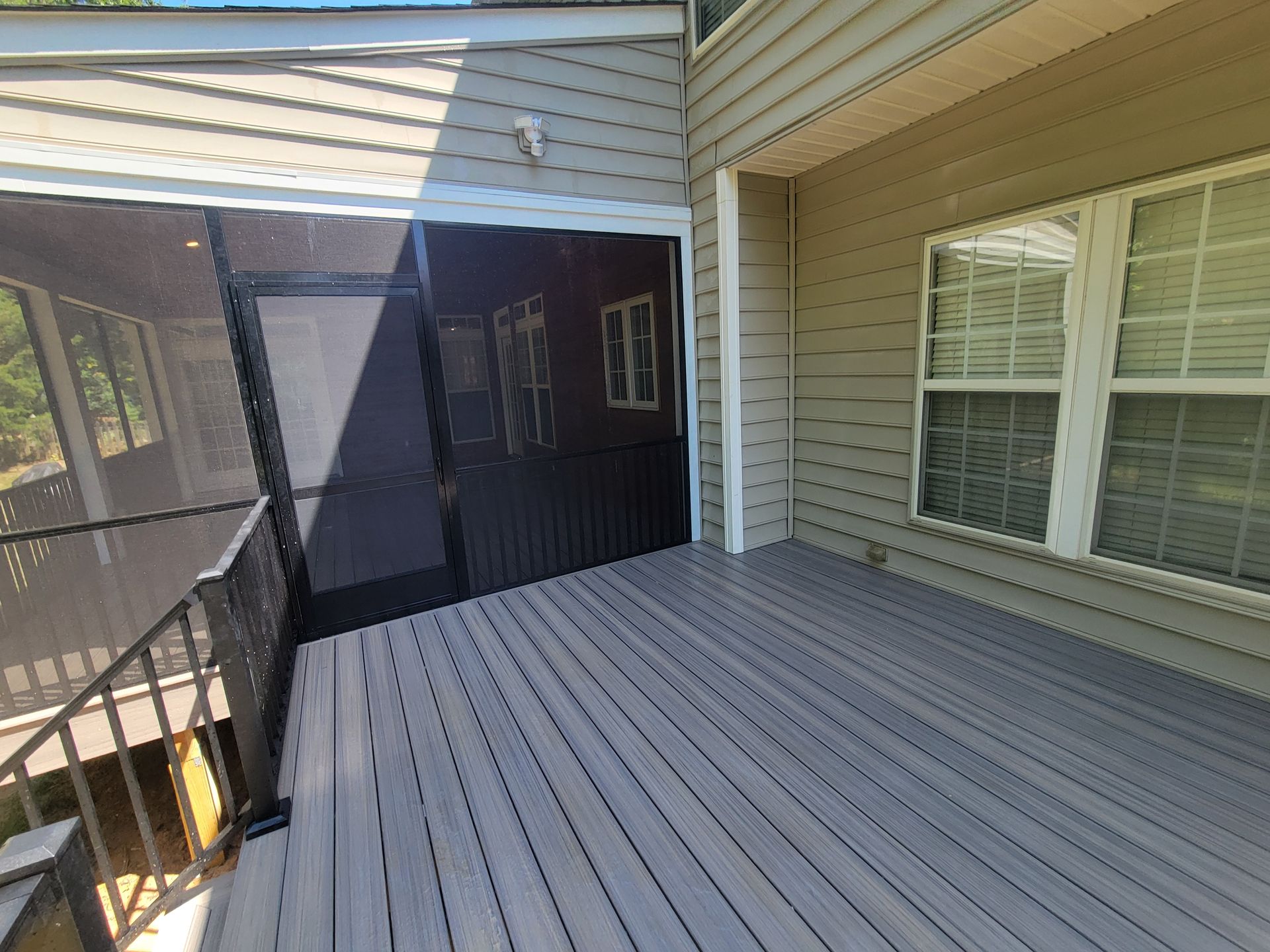 Covered patio with gray composite decking, beige siding, black screen door, and a window with white blinds.