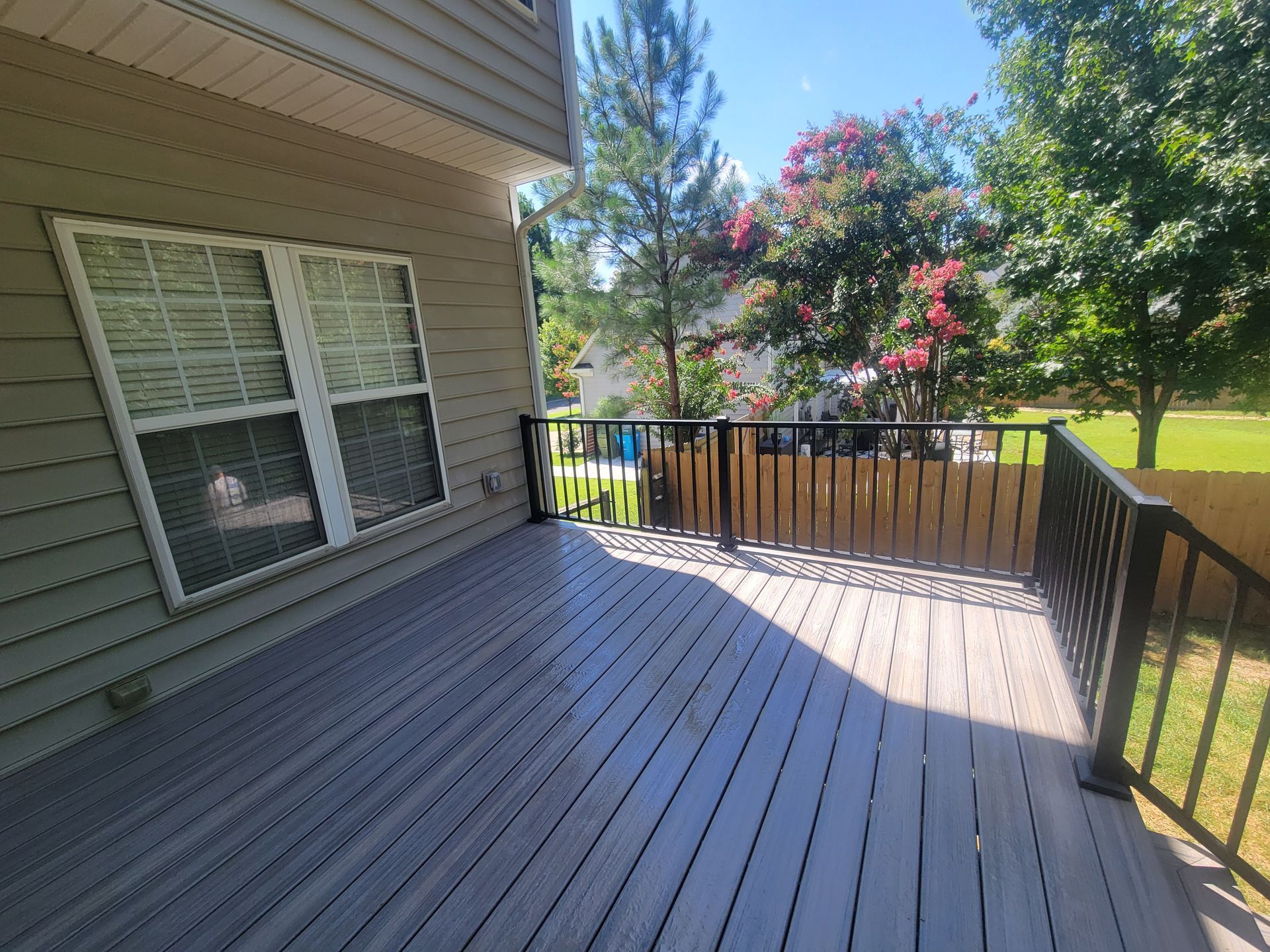 Back deck with gray wooden floor, black railing, house siding, and trees in the yard.
