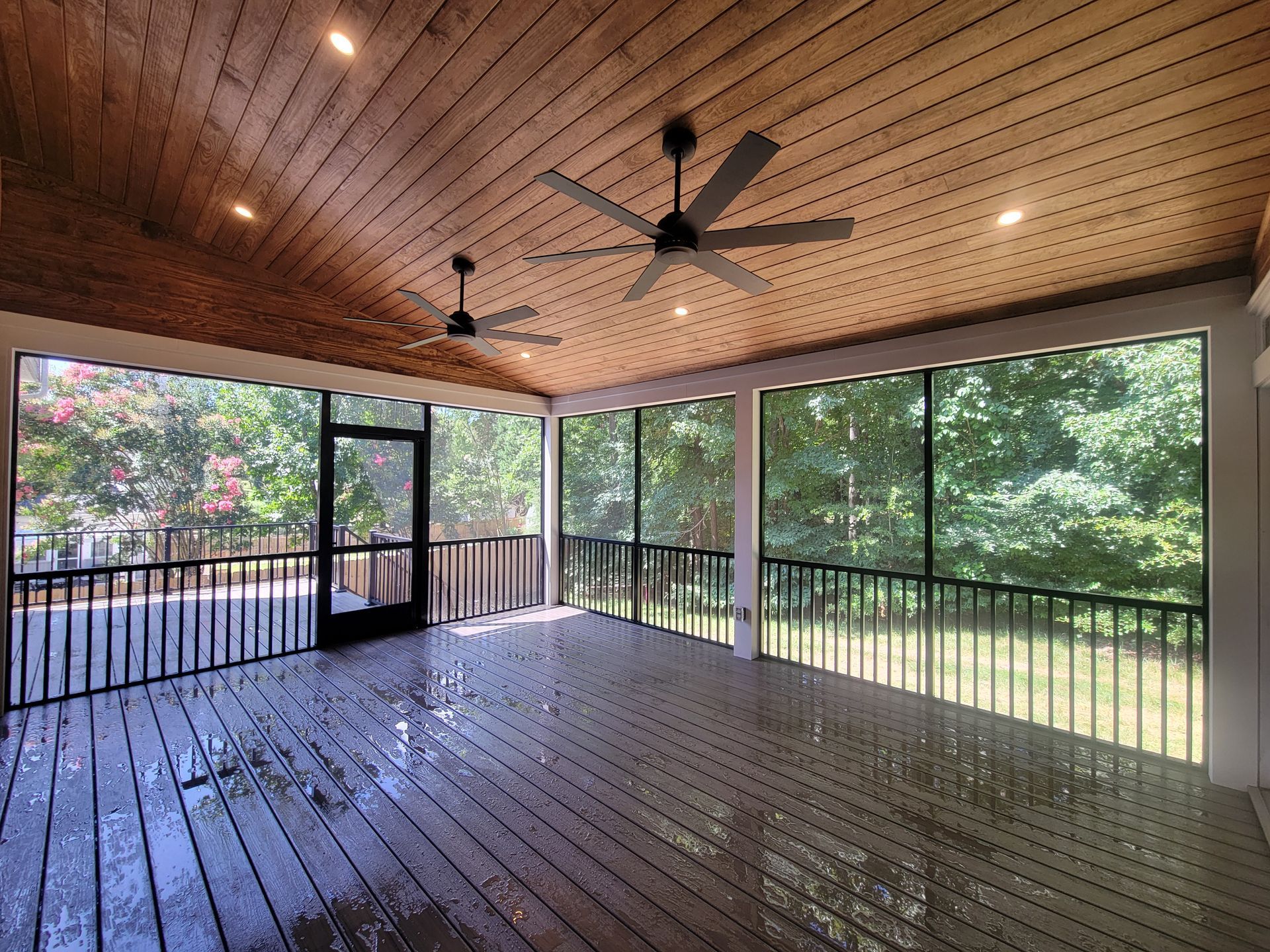 Screened porch with wood ceiling, ceiling fans, and wet floor overlooking green trees