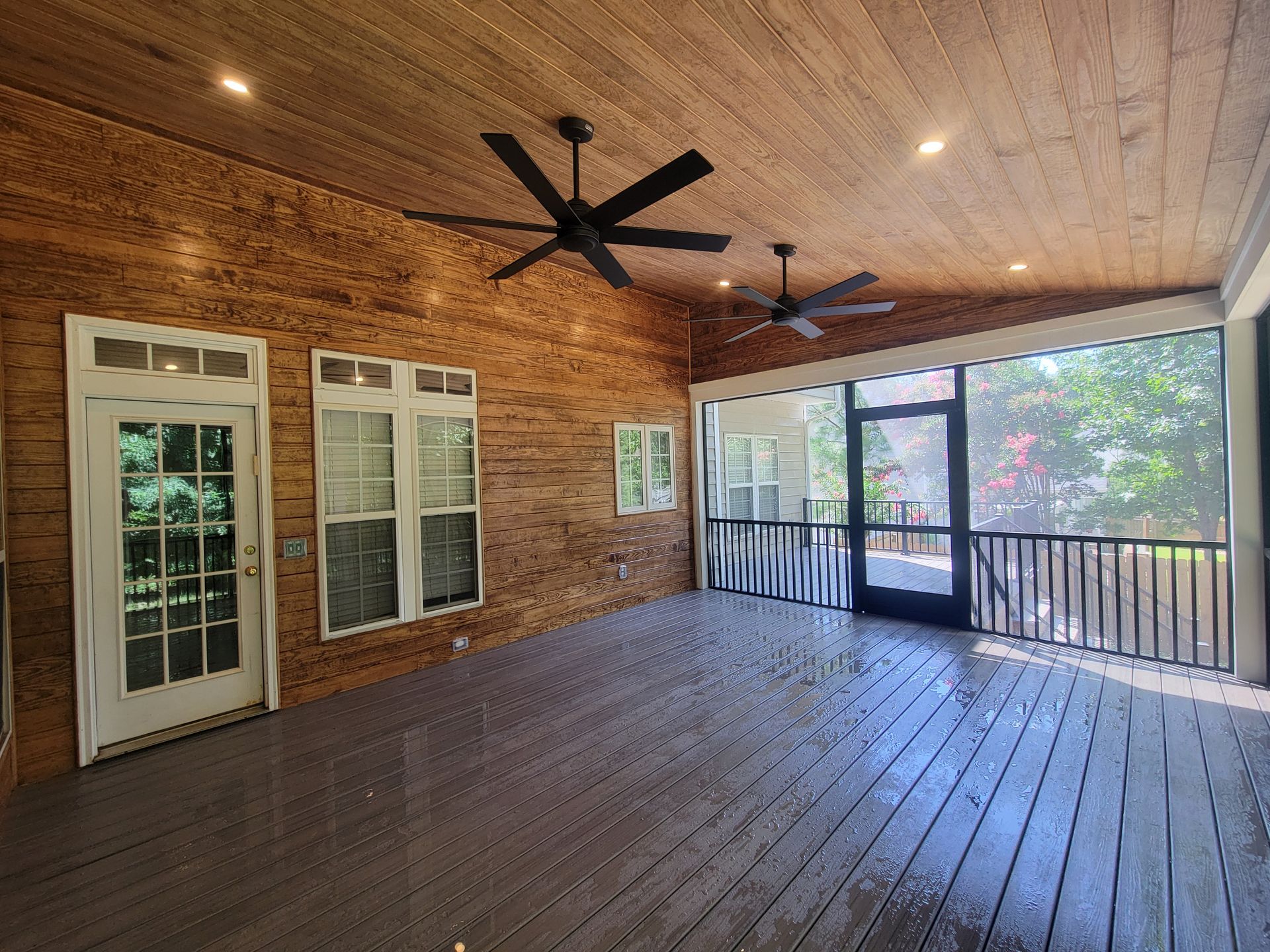 Covered brick porch with wooden ceiling, dark floor, ceiling fans, white doors, and railing overlooking trees.