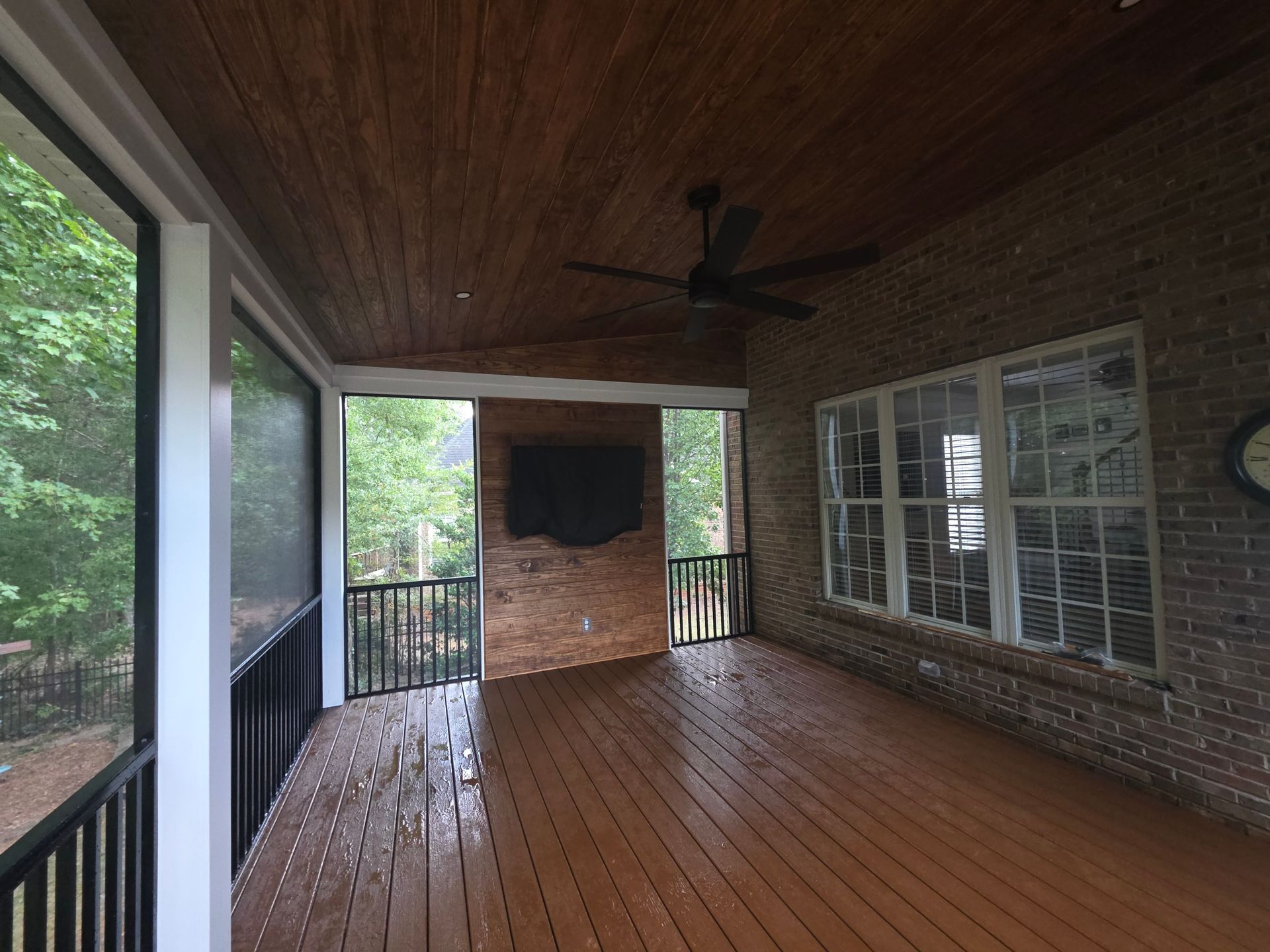 Covered wooden porch with brick wall, screened railing, ceiling fan, and large window looking out to greenery