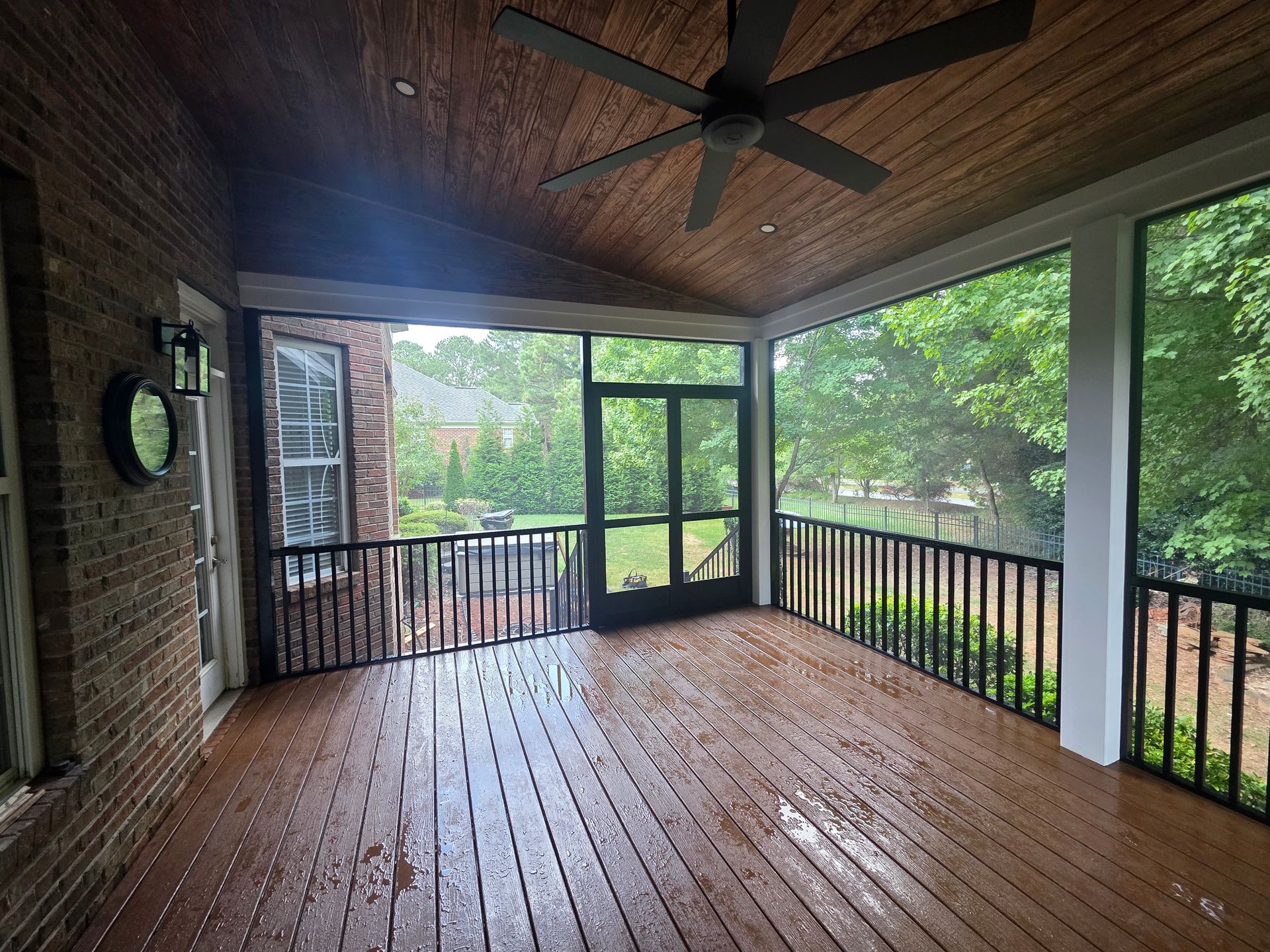 Covered wooden porch with screened windows, black railings, and a ceiling fan overlooking greenery.