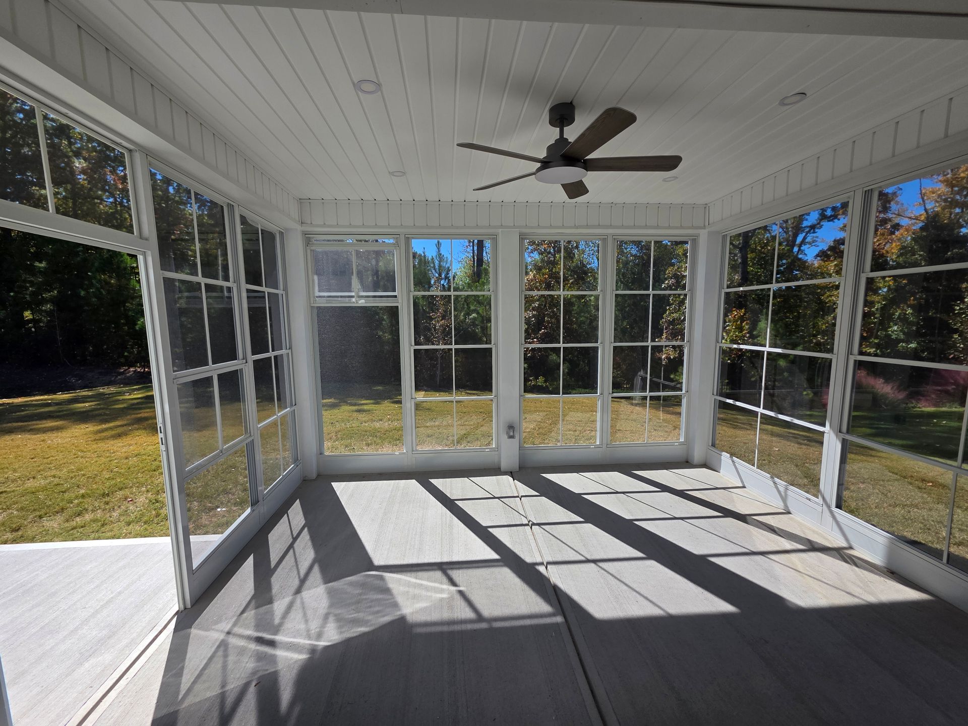 Sunlit screened porch with white ceiling, ceiling fan, and glass doors opening to a yard