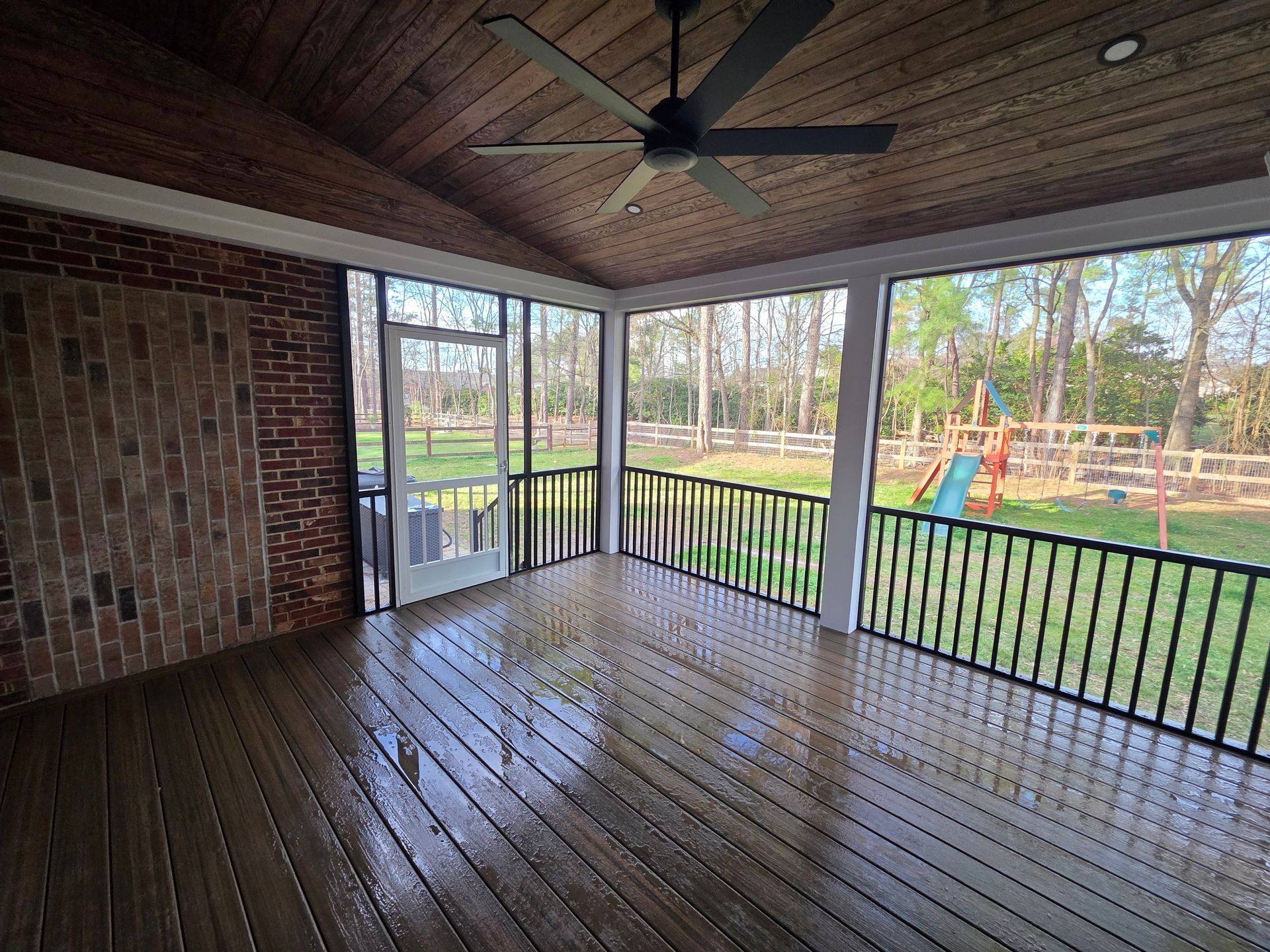 Covered wooden porch with black railings overlooking a yard and playground.