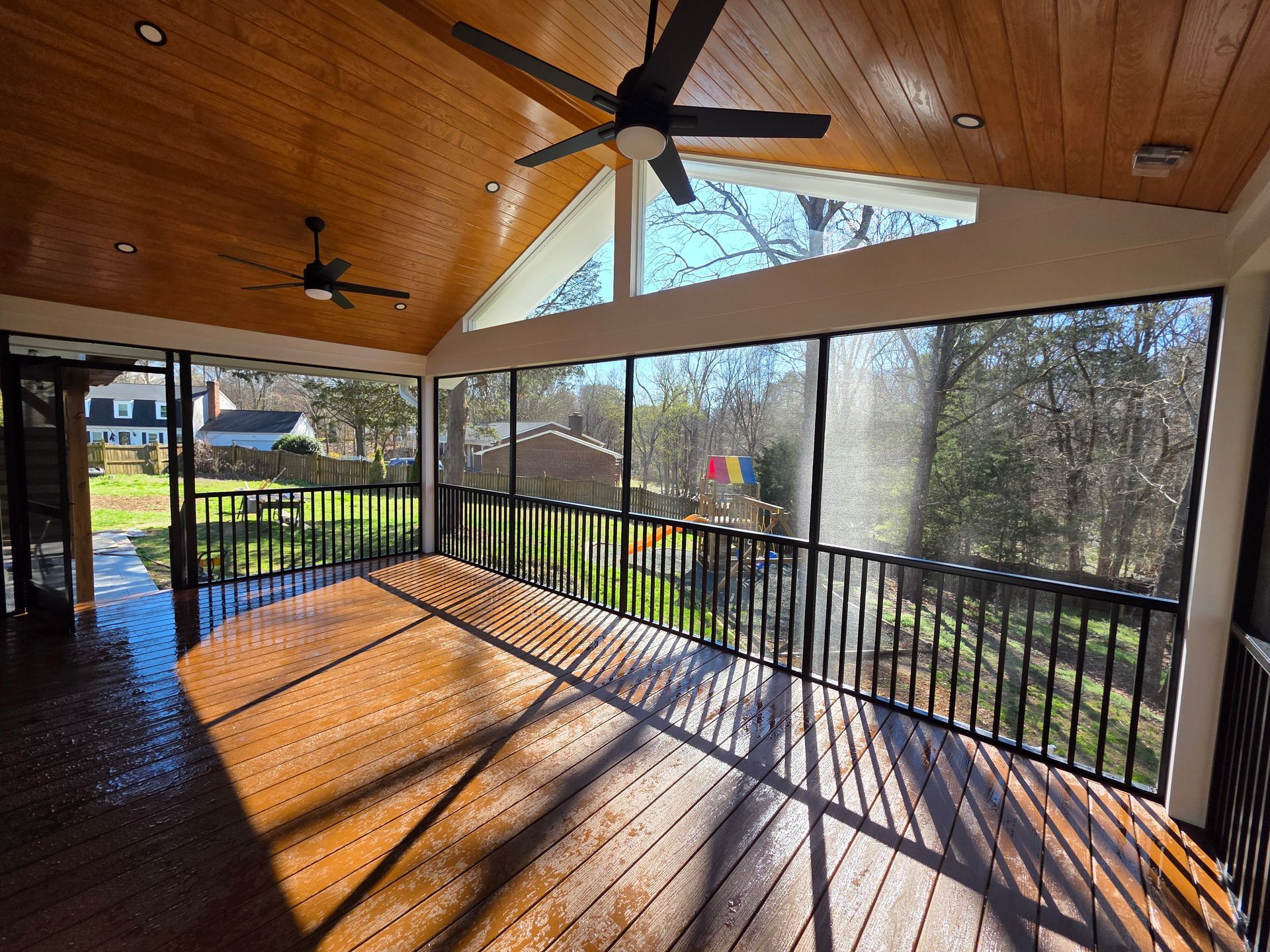 Covered wooden deck with black railing, ceiling fans, and sunlight streaming through screened openings.
