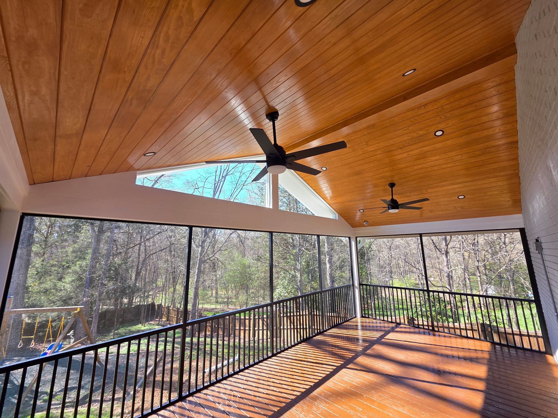 Covered porch with wood ceiling, ceiling fans, black railing, and sunlight casting shadows across the floor.