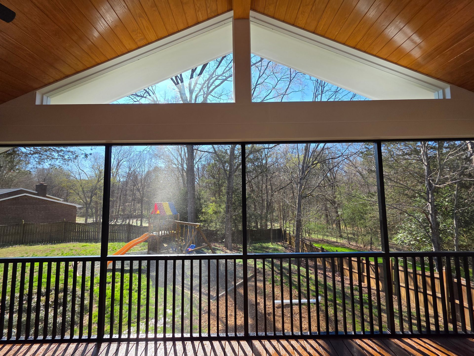 Covered porch view through black railing to trees and a sunny yard, with a peaked wood ceiling above.