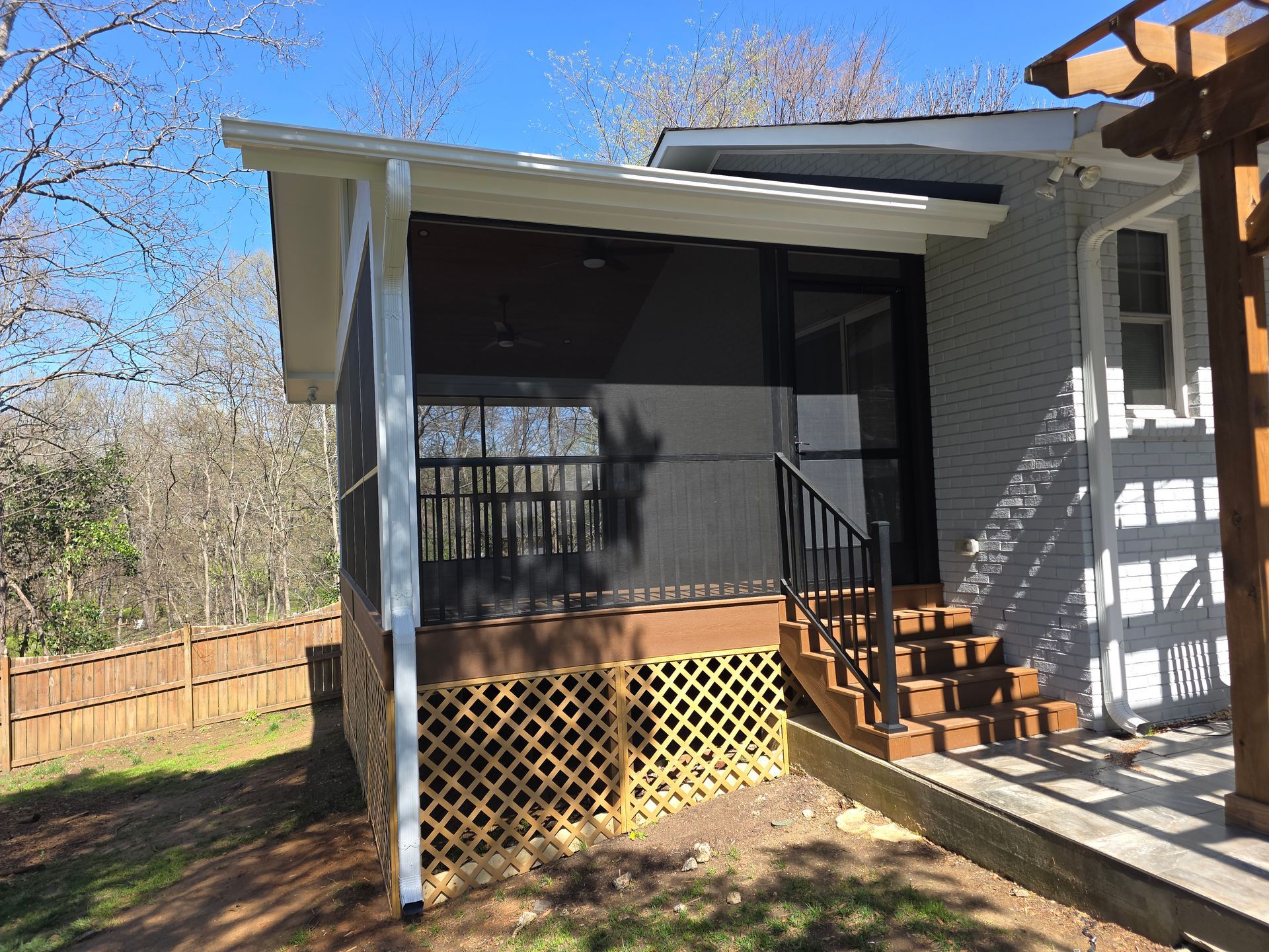 Covered porch with black railing and steps beside a gray house, with a fenced yard and trees beyond.