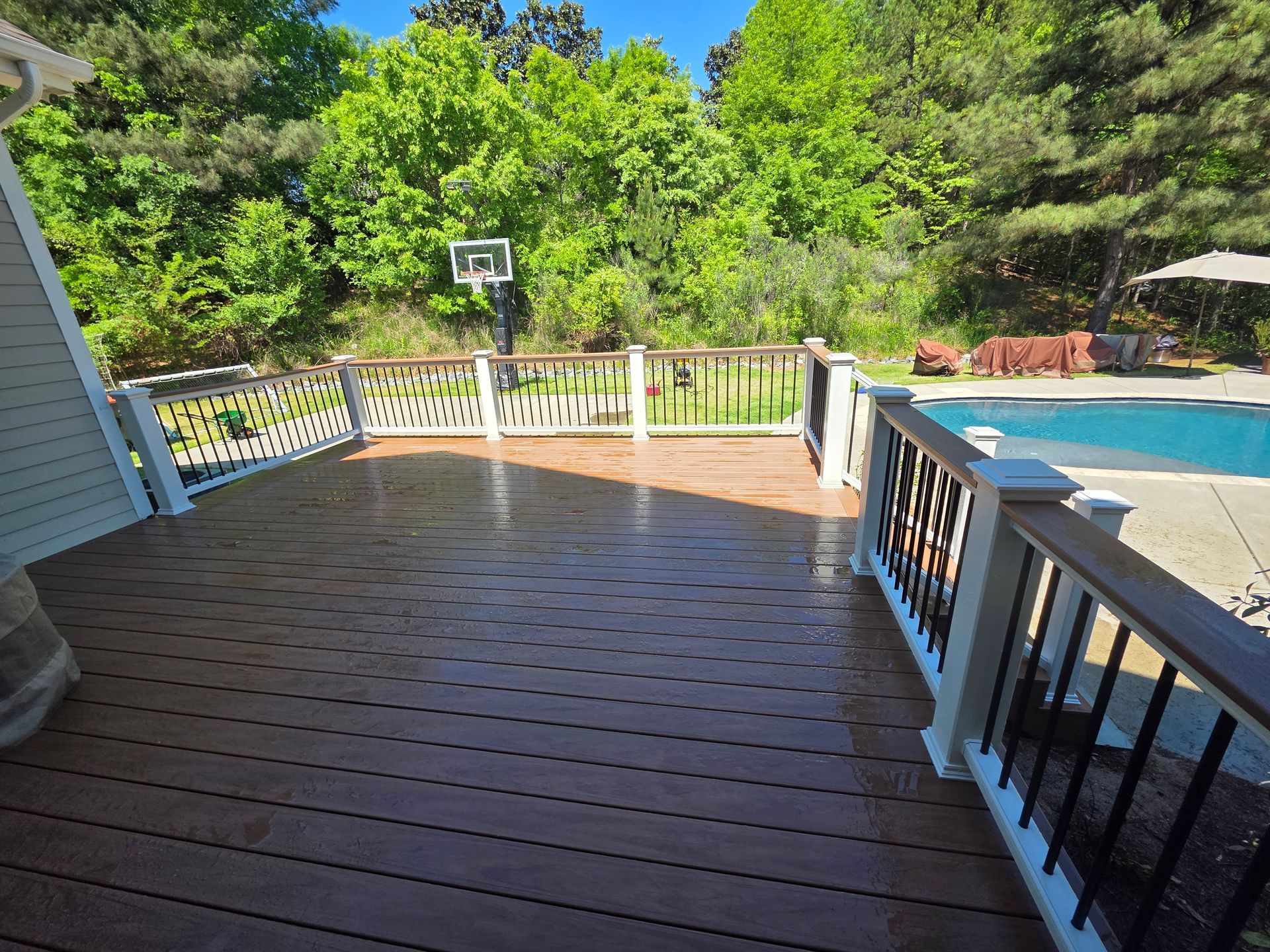 Wooden deck with railing overlooking a backyard pool and trees, with a basketball hoop in the distance