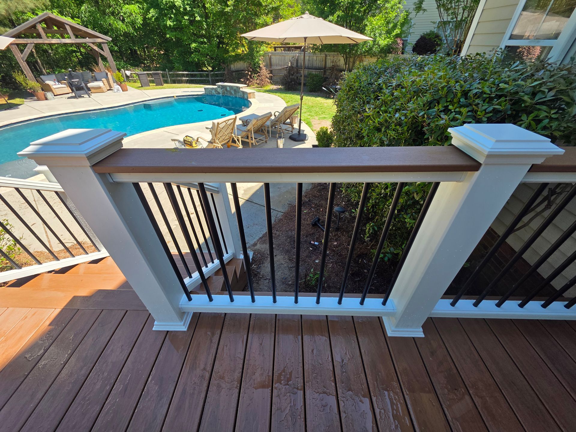 Wooden deck railing overlooking a backyard pool with umbrella and greenery.