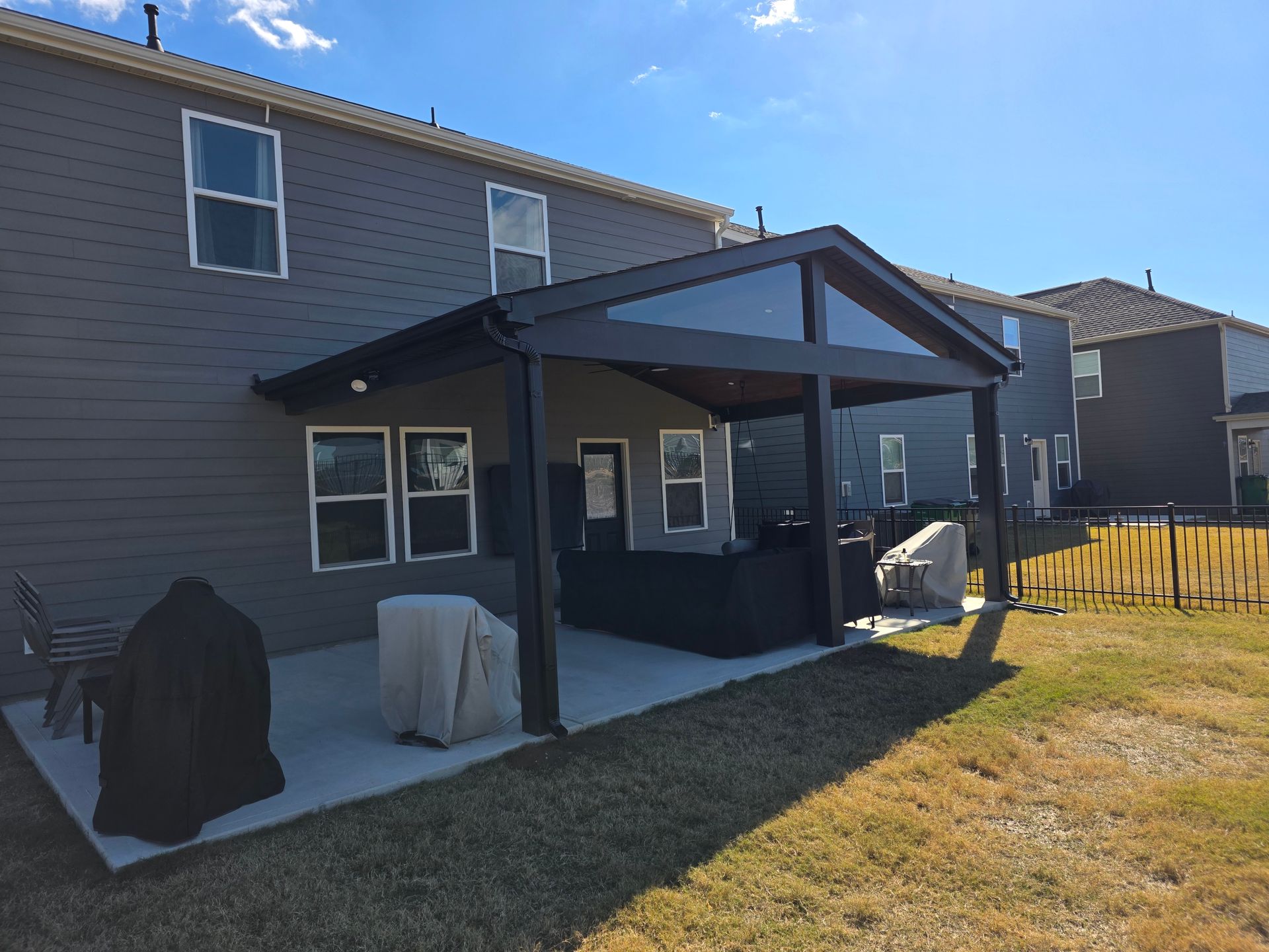 Back patio with dark covered pergola, grill, and patio furniture beside a two-story house and grassy yard