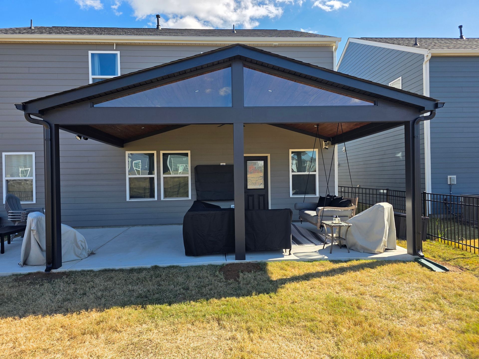 Covered patio with a dark metal roof, outdoor seating, and a grill in a backyard beside a house.