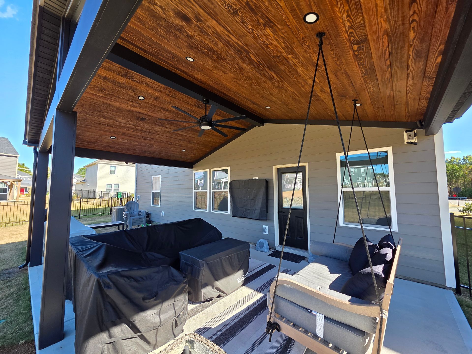Covered backyard patio with black outdoor furniture, wood ceiling, and sliding glass doors.