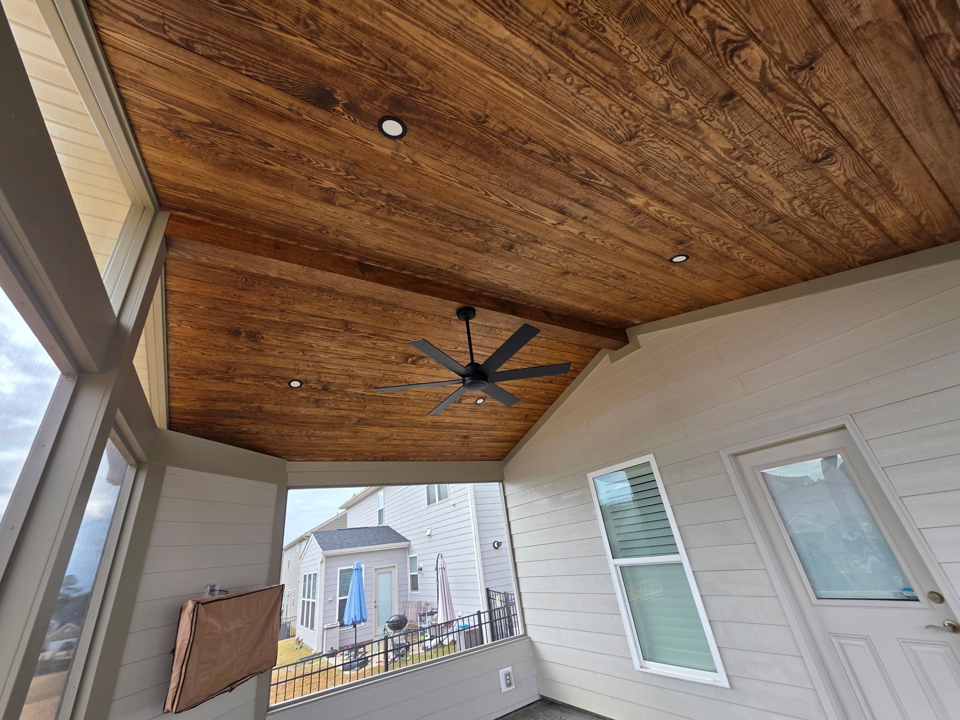 Covered porch with wood plank ceiling, ceiling fan, windows, and a view of neighboring houses.