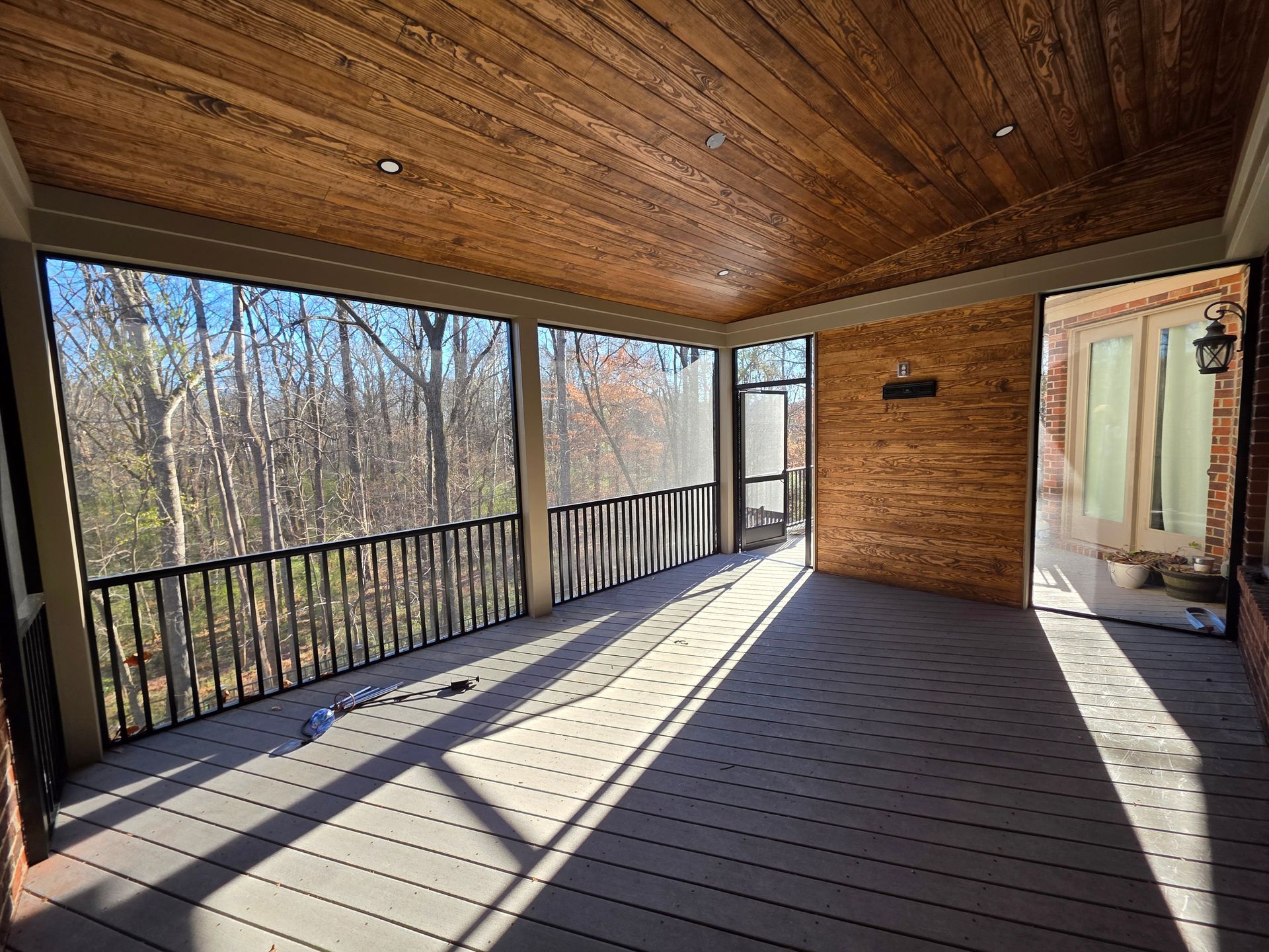 Covered wooden porch with slatted railings, brick wall, and sunlight casting long shadows over a wooded view