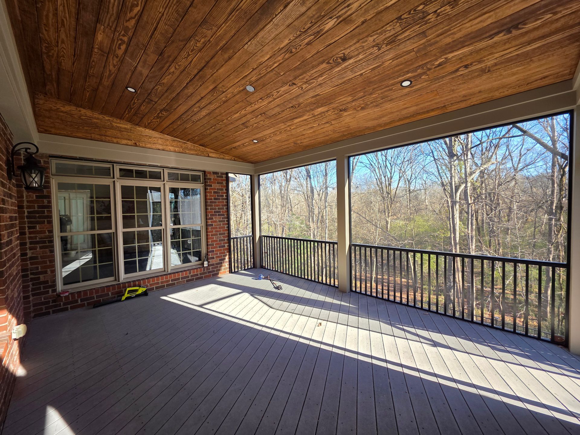 Covered wooden porch with black railings, brick wall, and trees beyond in bright sunlight
