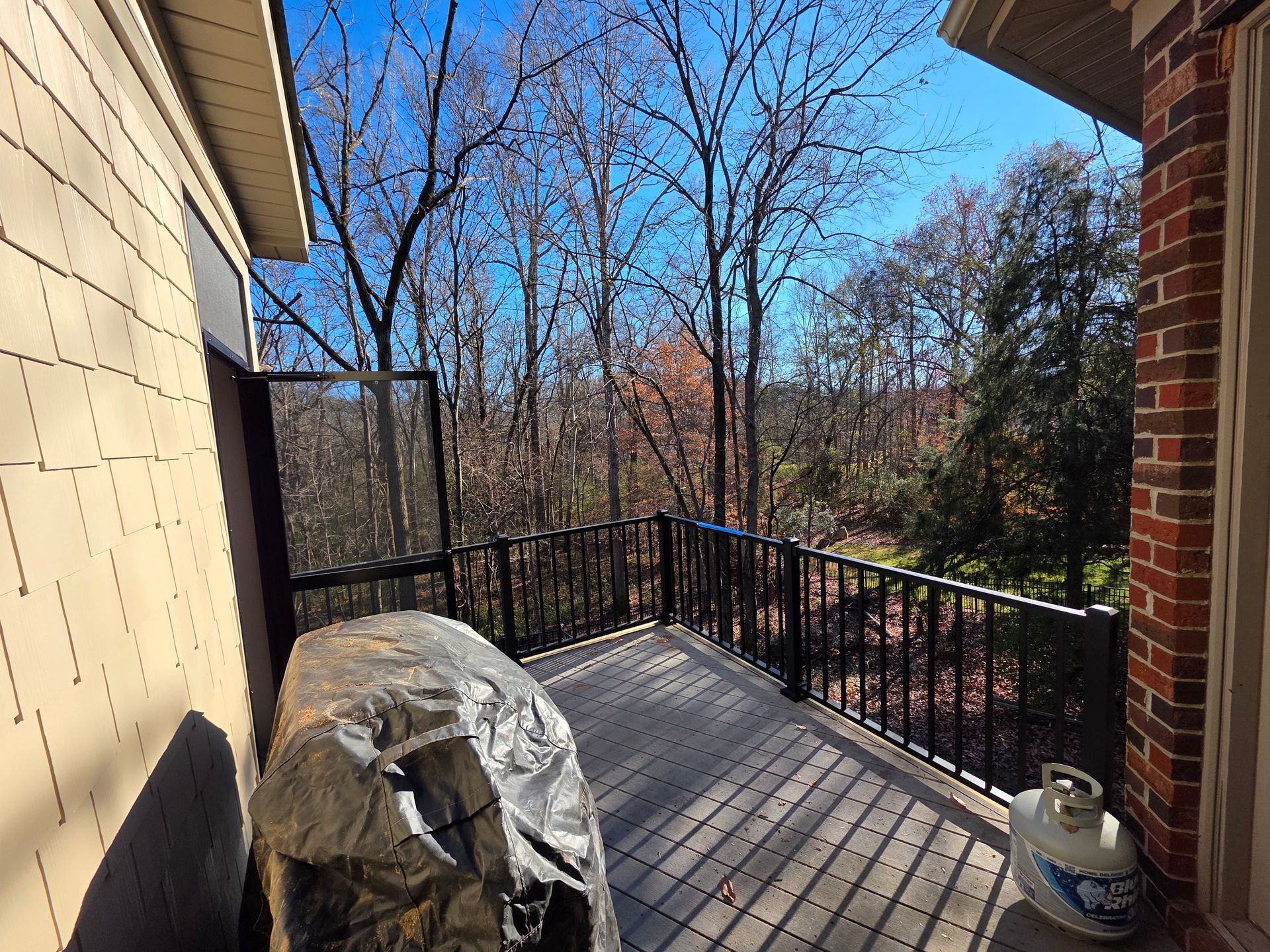 Sunlit balcony with black railing, covered grill, and trees beyond the view.