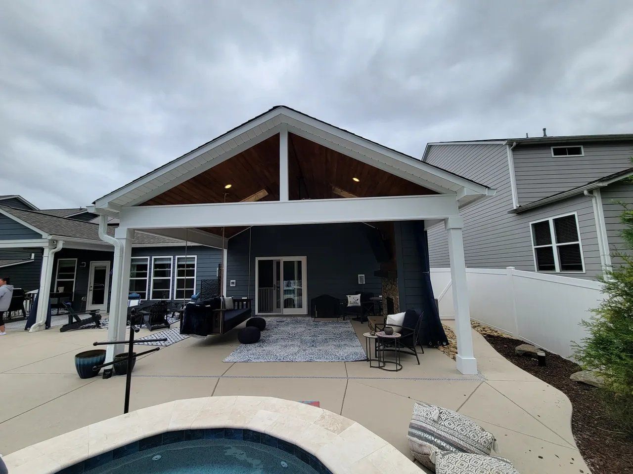 Covered patio with white columns, dark siding, seating, and a peaked wood ceiling in a backyard.