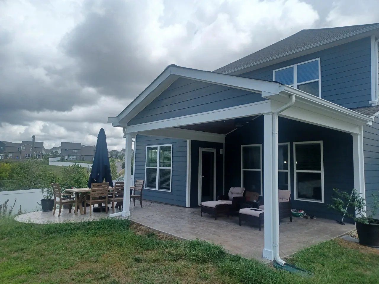 Back patio of a blue house with white trim, overlooking a lakeside yard under cloudy skies