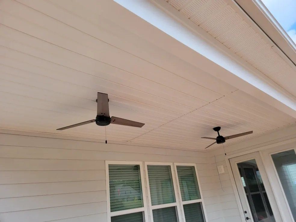 Covered patio ceiling with two black ceiling fans above white siding and windows