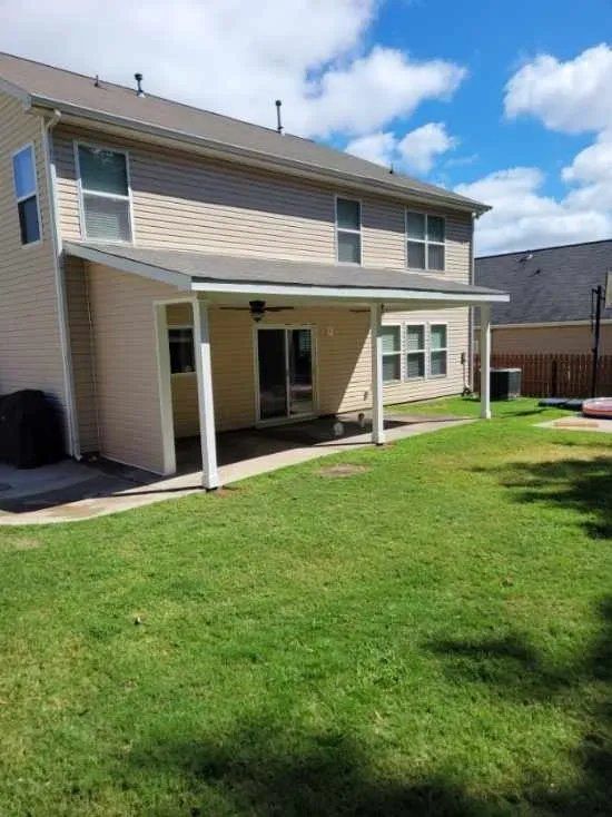 Two-story beige house with covered patio and white posts, overlooking a green backyard under a blue sky.