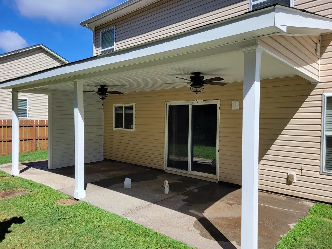 Covered patio with white columns, sliding glass doors, and beige siding beside a grassy yard.