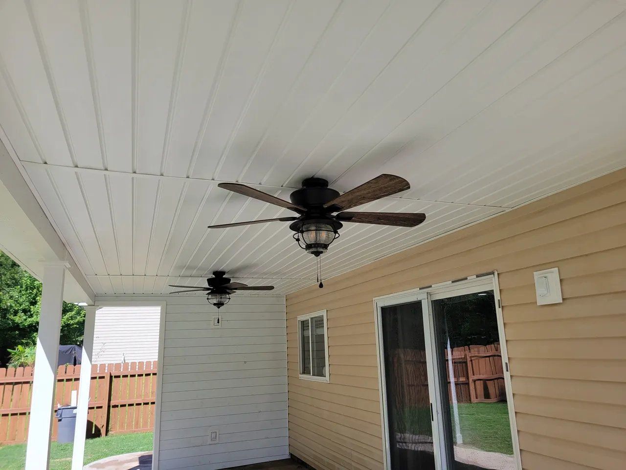 Covered patio with white beadboard ceiling, two ceiling fans, and beige siding beside a fenced yard.