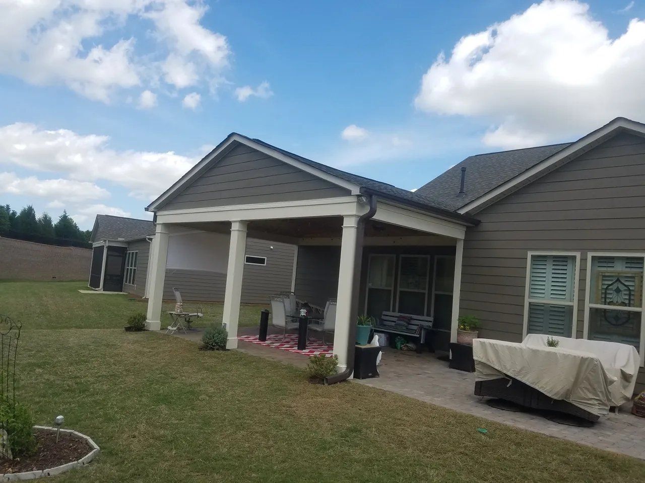 Covered patio on a suburban house with columns, potted plants, and a covered grill under a cloudy sky