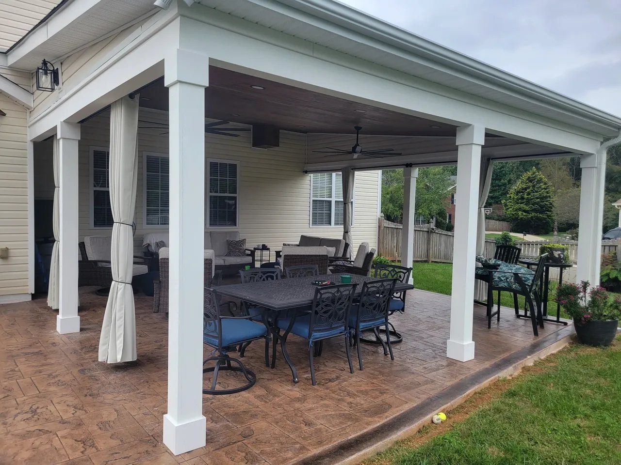 Covered patio with outdoor dining table and chairs, white columns, and tiled floor in a backyard.