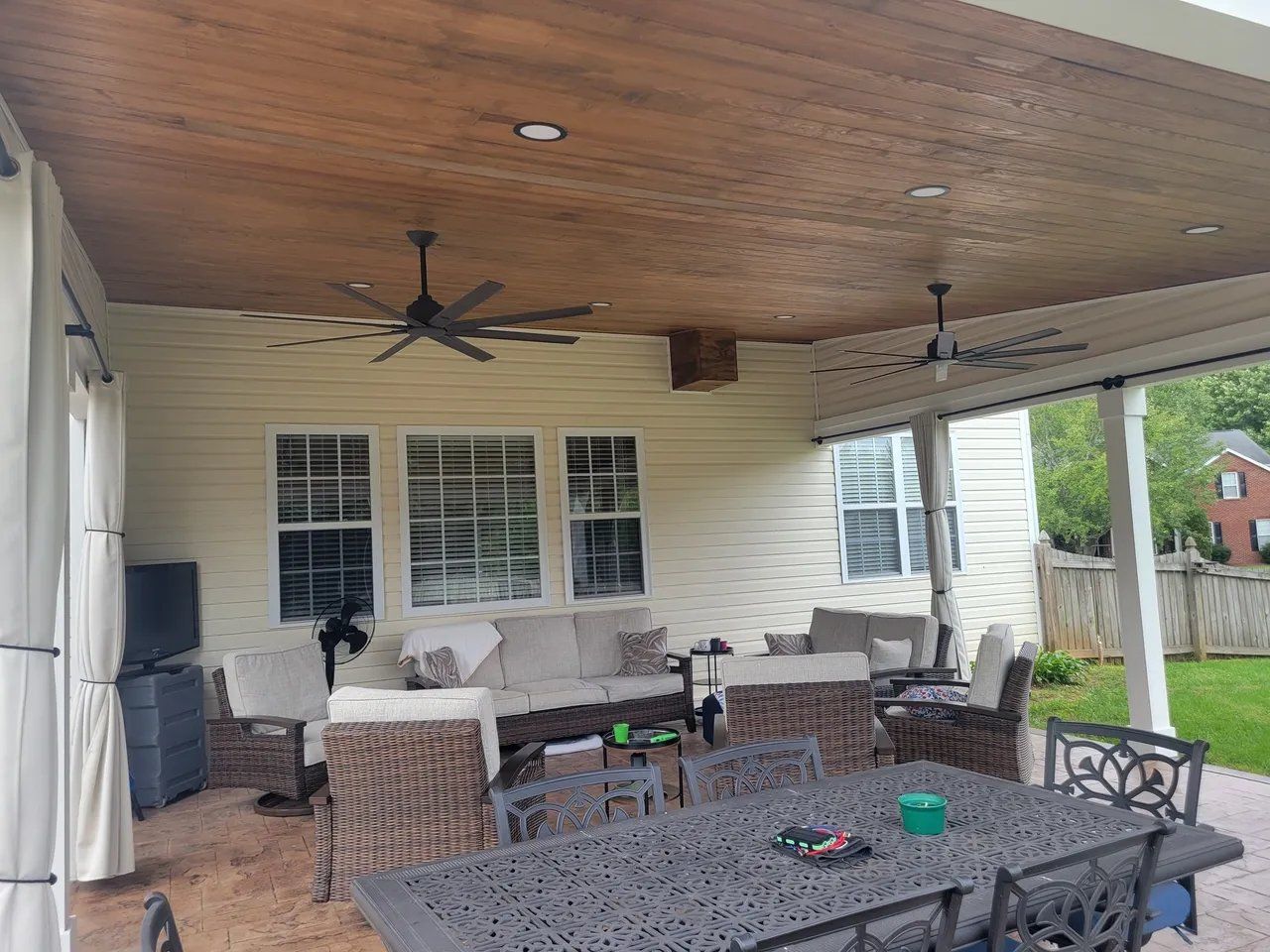 Covered patio with wicker seating, ceiling fans, and a dining table beside a grassy yard.