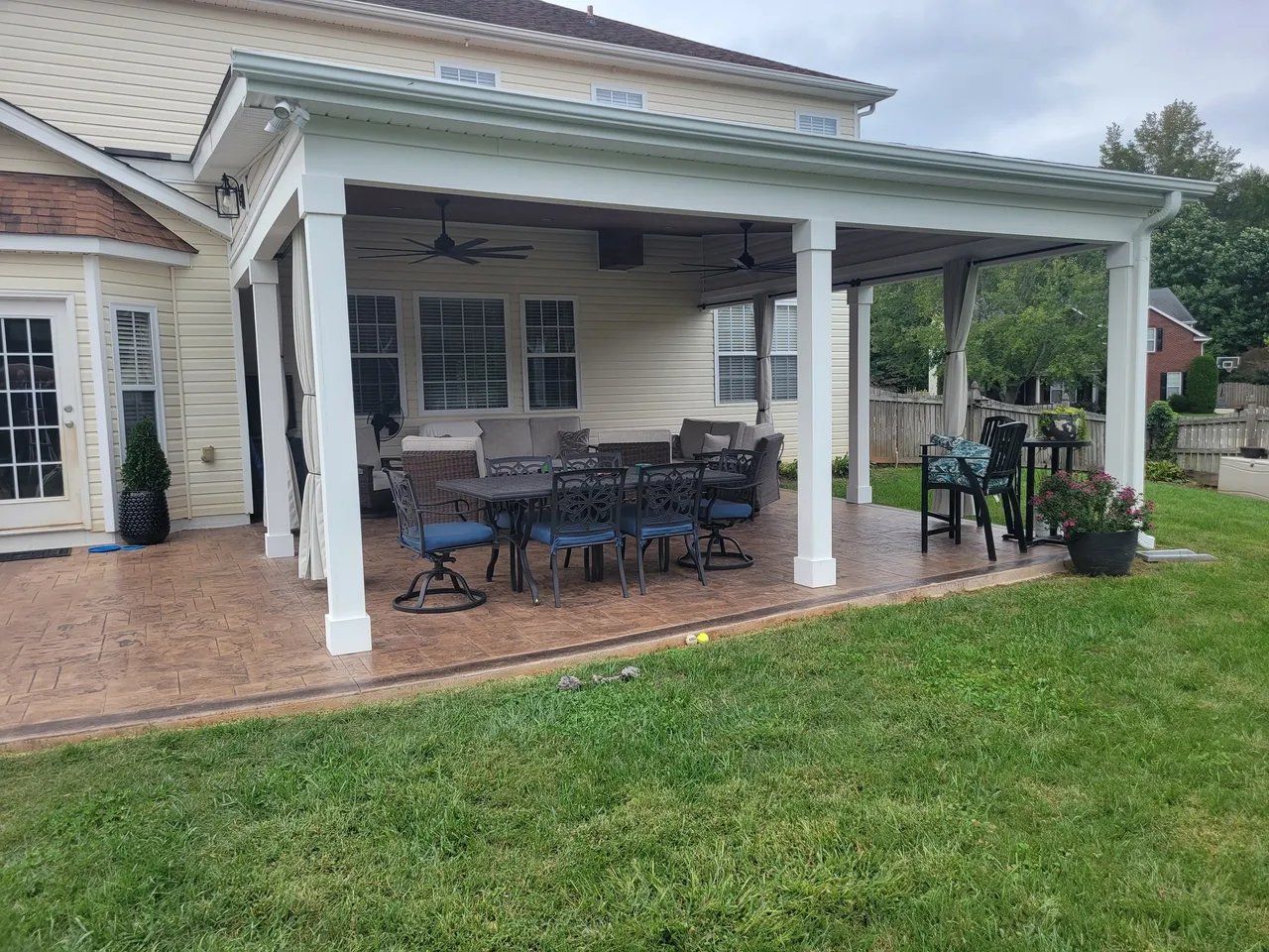 Covered patio with white columns, outdoor dining table, and chairs beside a house and green lawn