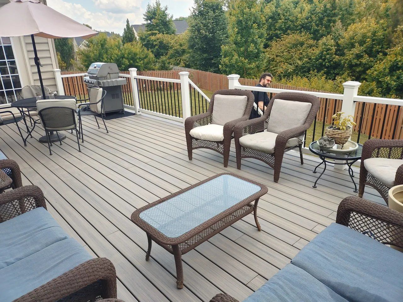 Patio deck with outdoor seating, glass coffee table, grill, and trees beyond the railing