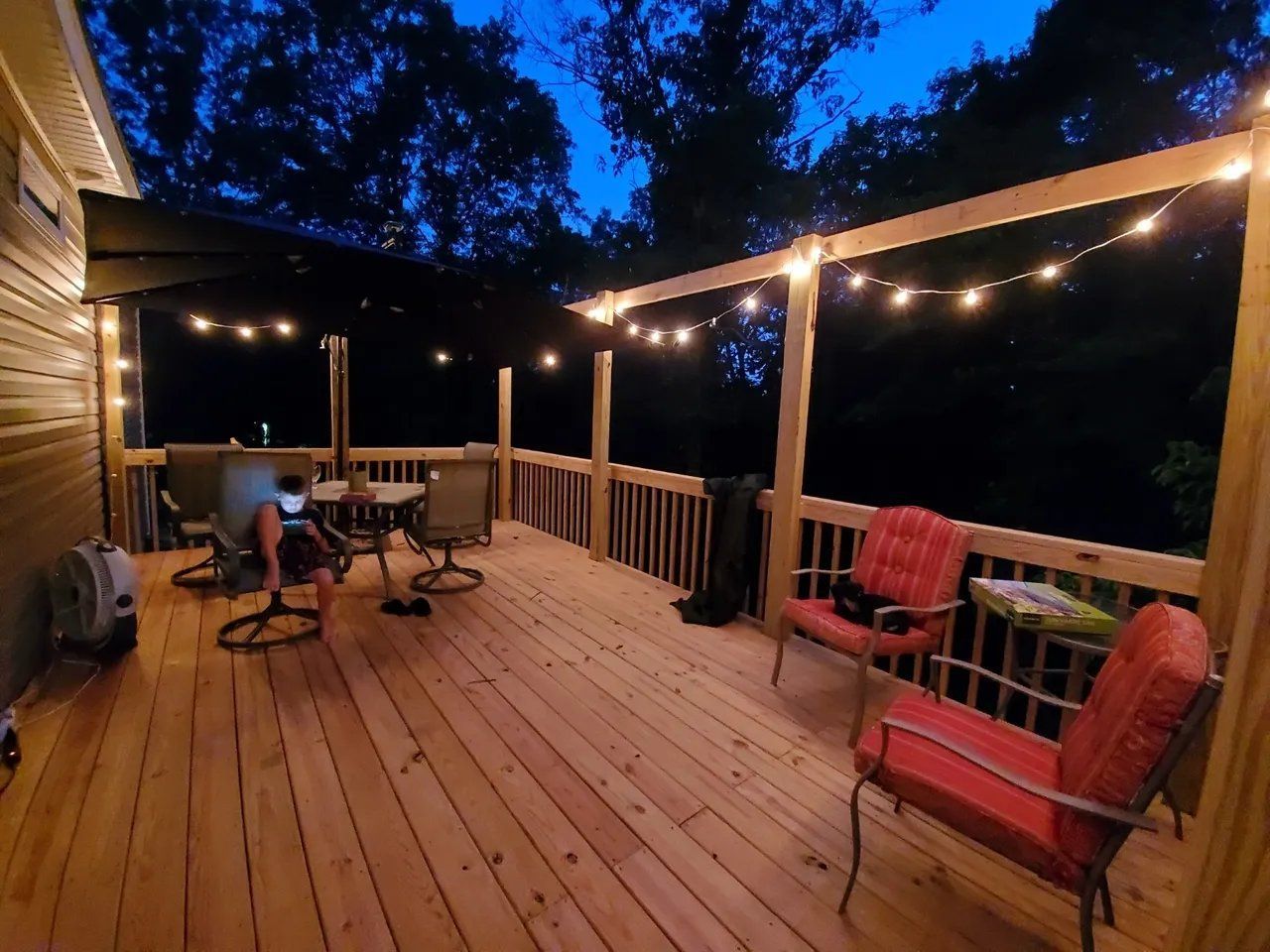 Covered wooden deck at dusk with string lights, patio chairs, and a small table overlooking dark trees.