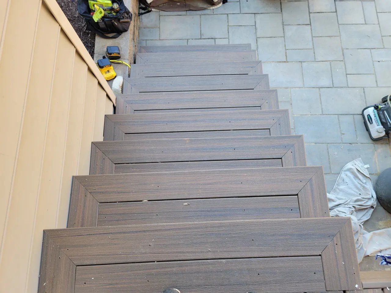 Wooden stairs beside a beige house leading down to a paved patio with tools and supplies nearby