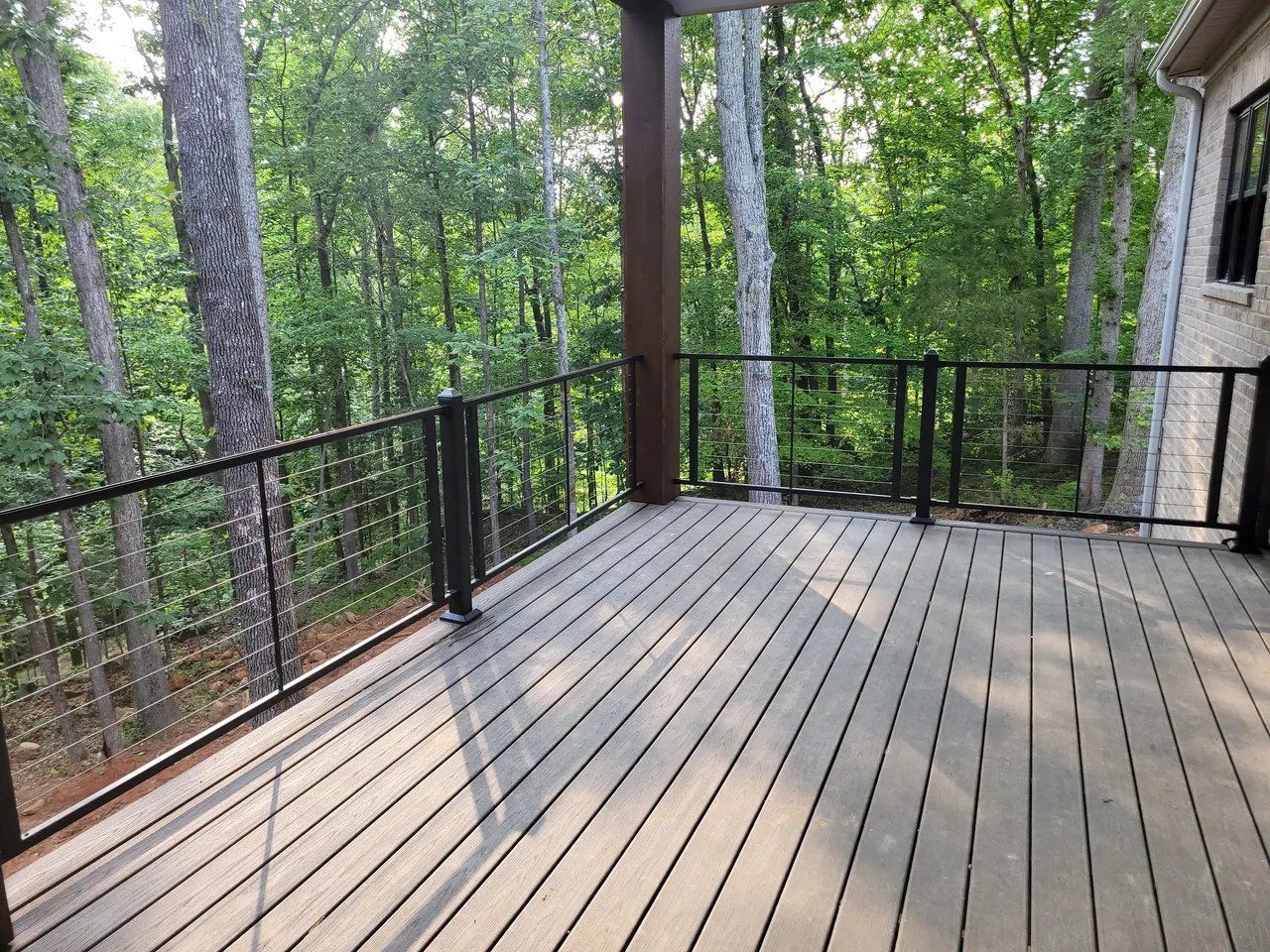 Wooden deck with black railing overlooking a wooded forest beside a stone house