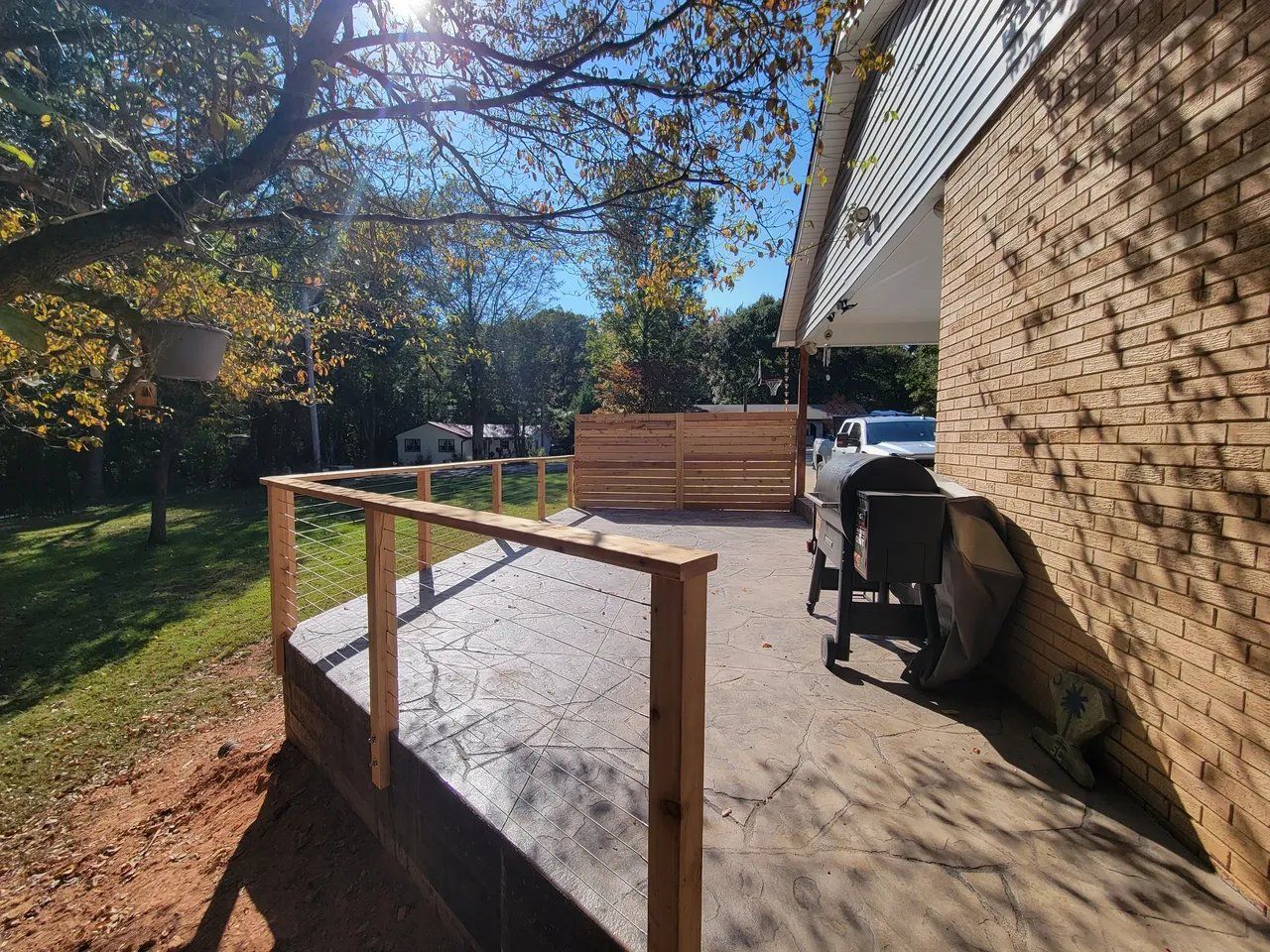 Backyard patio with ramp railing, brick house wall, and grill in sunlight.