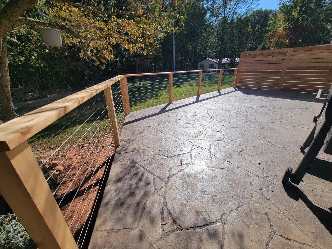 Wooden deck with wire railing and sunlit concrete path overlooking trees and hillside outdoors