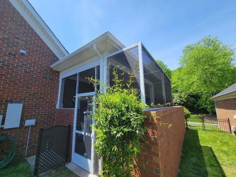 Brick house sunroom with screened windows, white roof, and a green vine-covered fence in a sunny yard