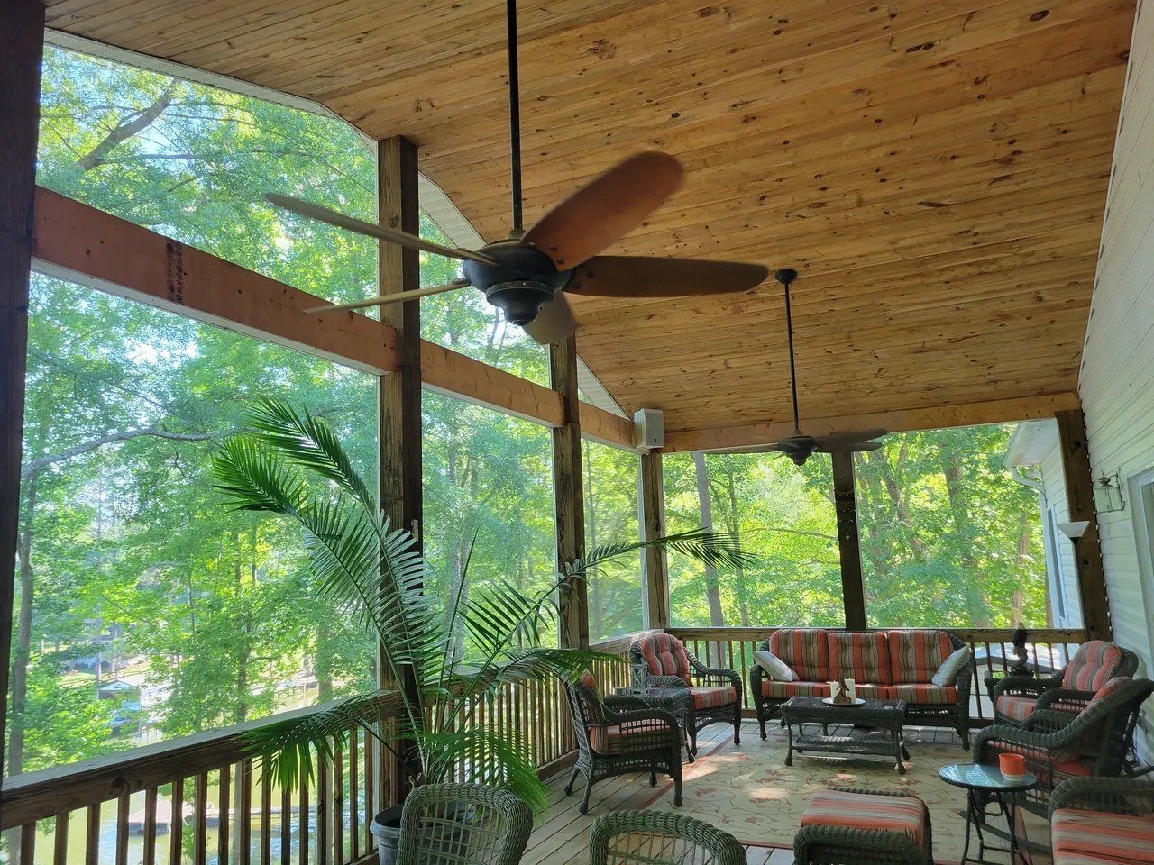 Covered porch with wooden ceiling, ceiling fan, wicker seating, and lush green view outside