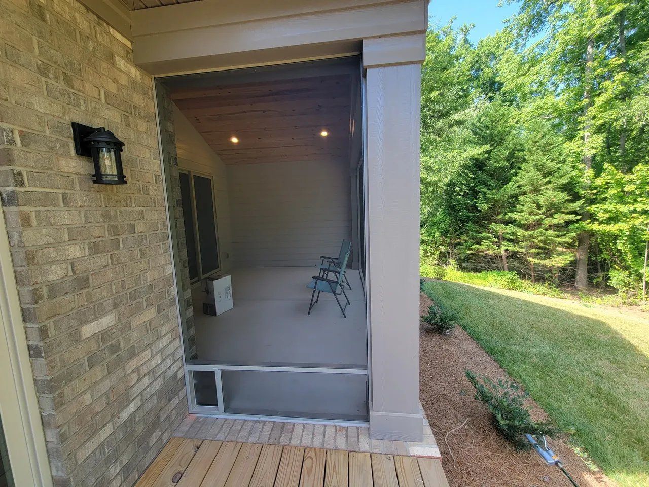 Covered brick porch with a chair, white columns, and a grassy yard beside a tree-lined driveway.